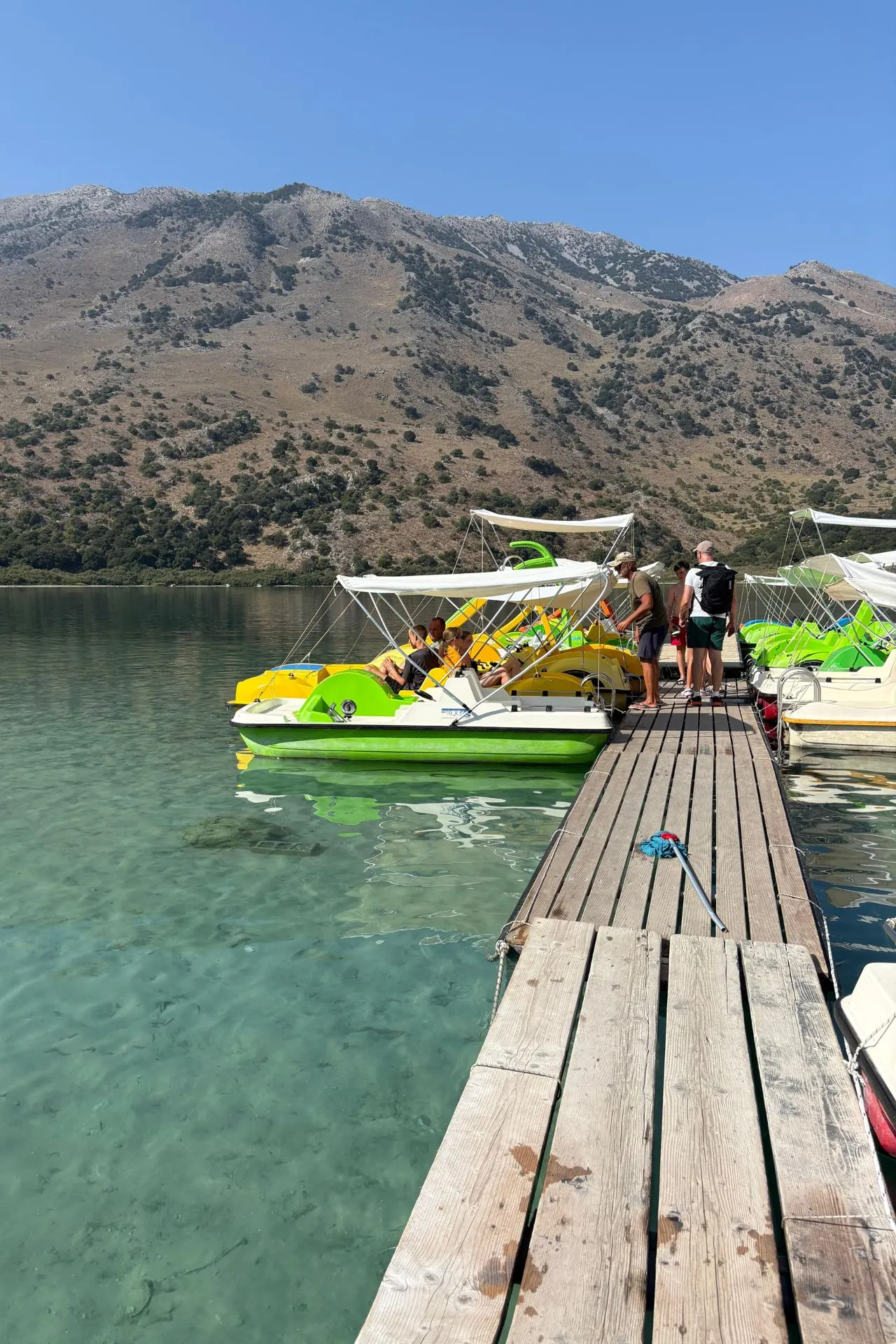 Groen-gele waterfietsen aangemeerd aan een houten steiger op helderblauw water, omringd door bergen onder een strakblauwe lucht