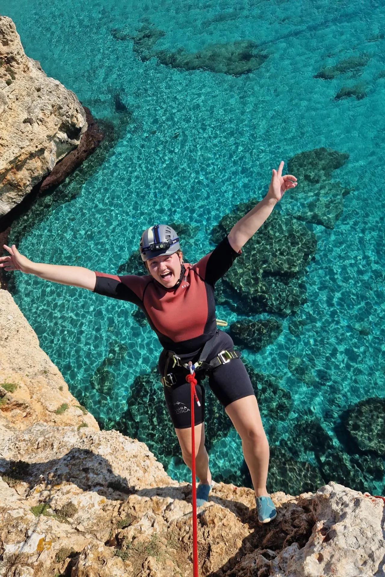 Persoon in wetsuit en klimgordel staat op een rots aan helderblauw water op Mallorca, klaar om te springen of te balanceren tijdens een avontuurlijke excursie.
