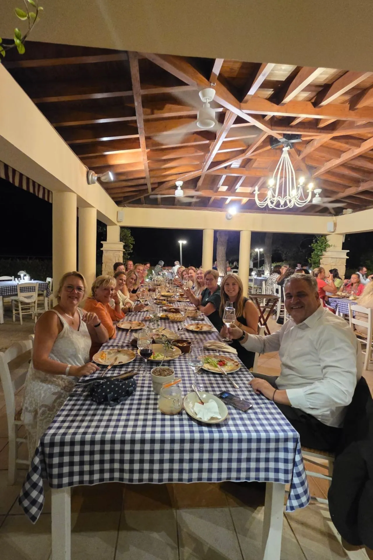 Grote groep mensen aan een lange tafel met blauw-wit geruite tafelkleden, gedekte met borden, glazen en traditionele gerechten, onder een houten veranda met verlichting.