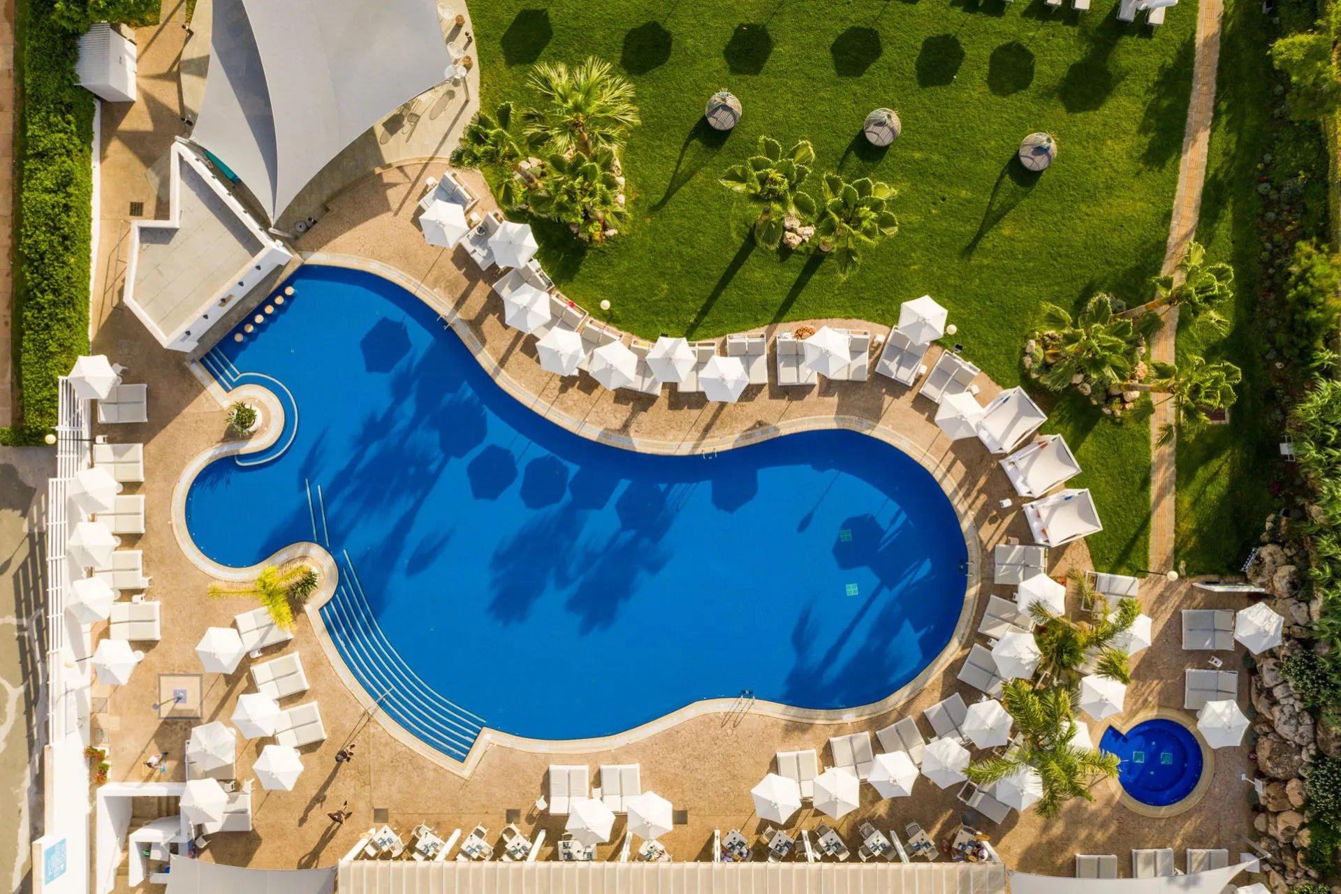 Luchtfoto van een Atlantica resort met een groot niervormig zwembad omgeven door witte ligbedden en parasols. De schaduw van palmbomen valt over het felblauwe water, terwijl een jacuzzi en een verzorgde tuin de luxe uitstraling versterken.
