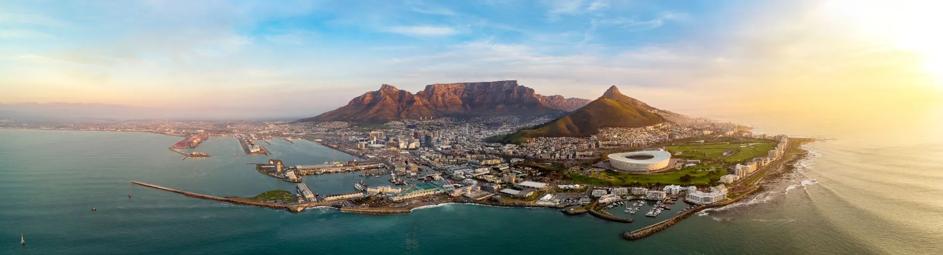 Panoramisch uitzicht op Kaapstad in Zuid-Afrika met de iconische Tafelberg op de achtergrond, de Atlantische Oceaan aan de linkerkant en het moderne stadion prominent in beeld. De stad strekt zich uit langs de kustlijn met een mix van stedelijke gebouwen, groene parken en havens, verlicht door een warme zonsondergang. Ideaal voor SEO-thema’s zoals reizen naar Zuid-Afrika, vakantie in Kaapstad, Tafelberg bezoeken, kuststeden ontdekken en avontuurlijke bestemmingen in Afrika.