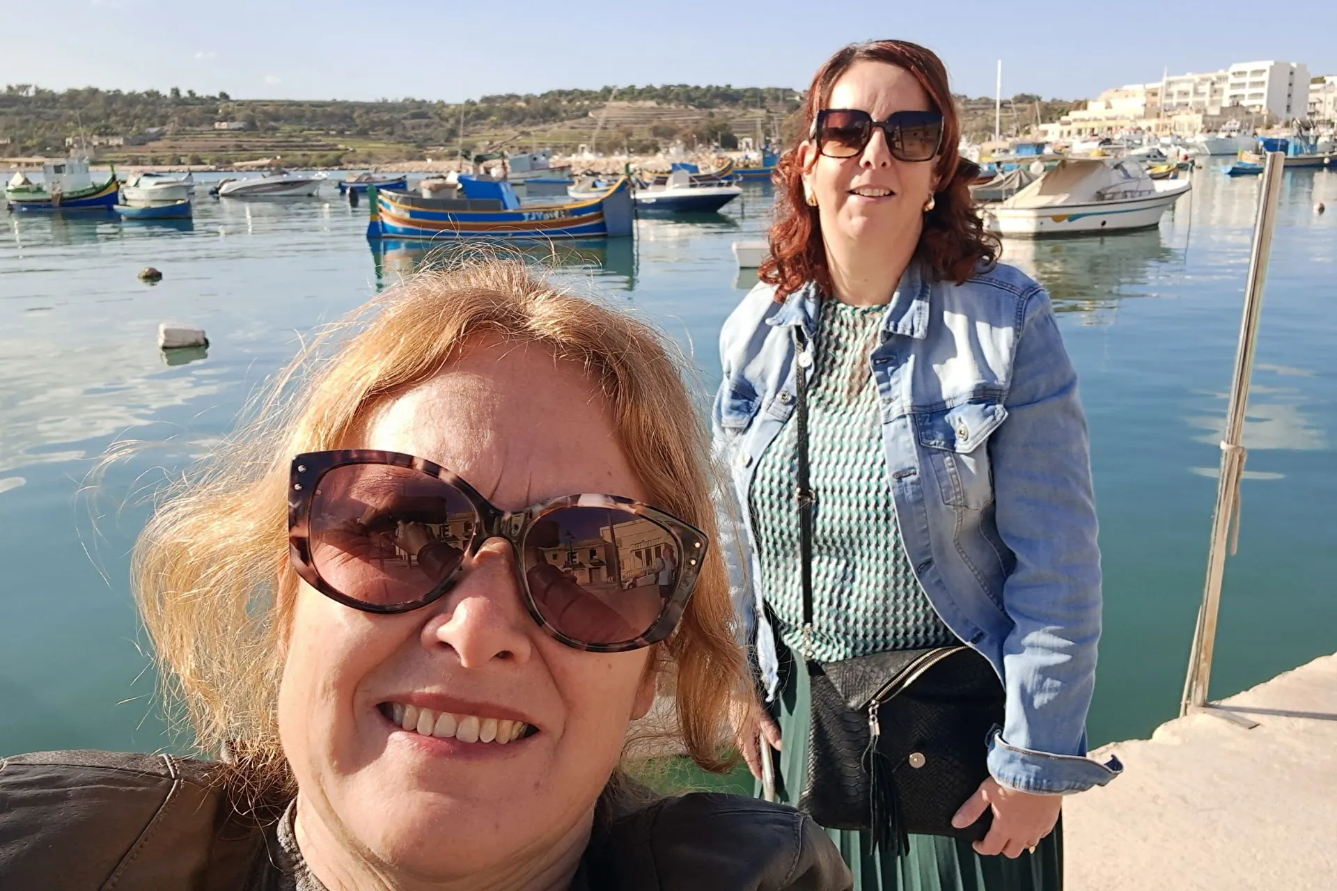Reisadviseurs Joyce en Karin op Malta, aan het water met bootjes op de achtergrond.