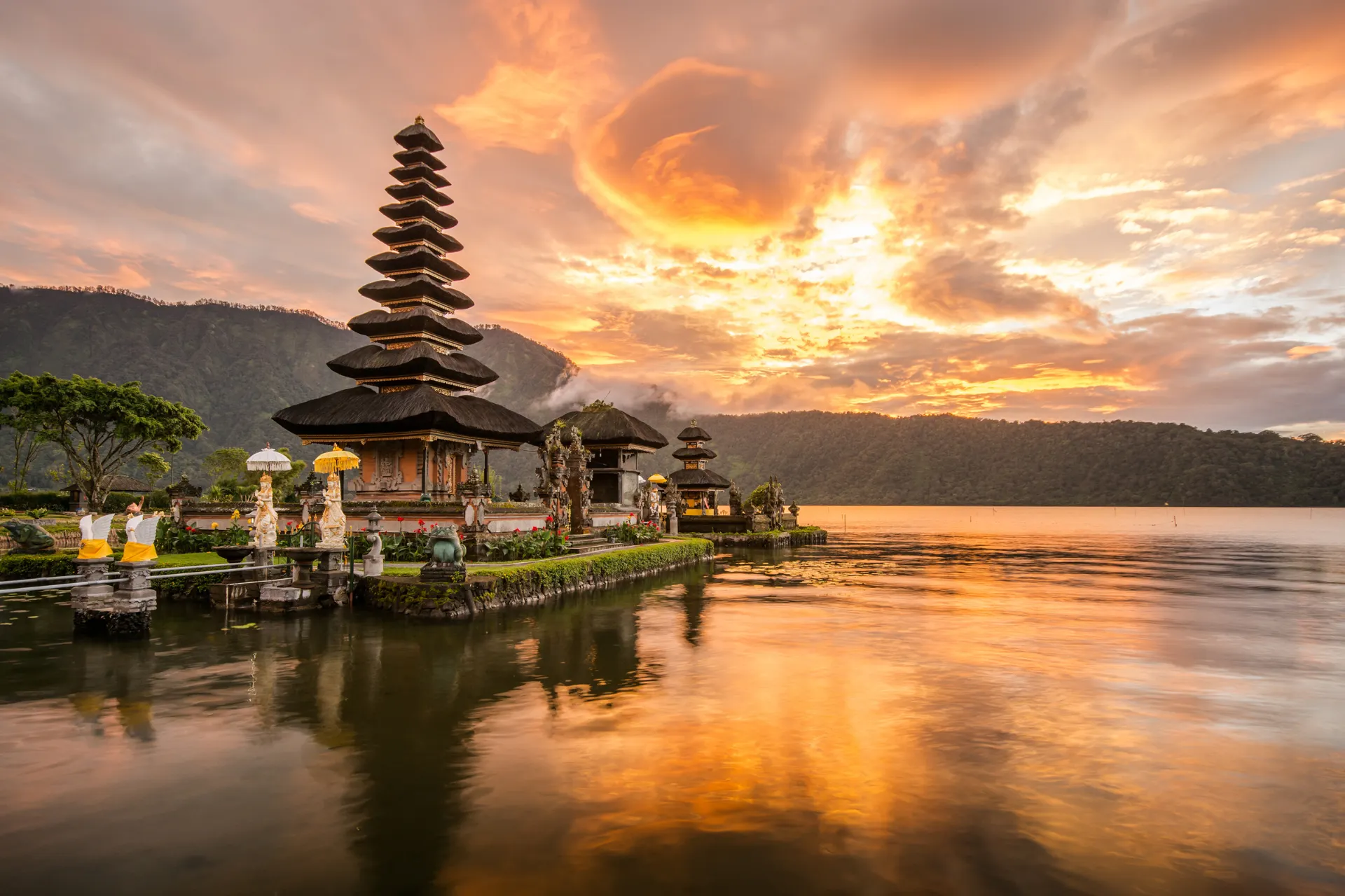 "Traditionele Balinese tempel aan het water bij zonsondergang, omringd door bergen en weelderige tropische natuur. Spectaculair uitzicht met gouden lucht en serene reflecties in het meer, ideaal voor cultuurreizen, fotografie en spirituele ervaringen. Ontdek authentieke architectuur, exotische landschappen en unieke reisbestemmingen in Indonesië. Perfect voor liefhebbers van natuur, erfgoed en ontspanning."
