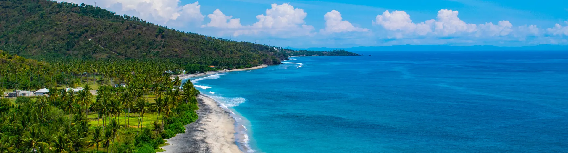 Zee in Indonesië met helder water rechts van een zandstrand, omringd door groene bomen.