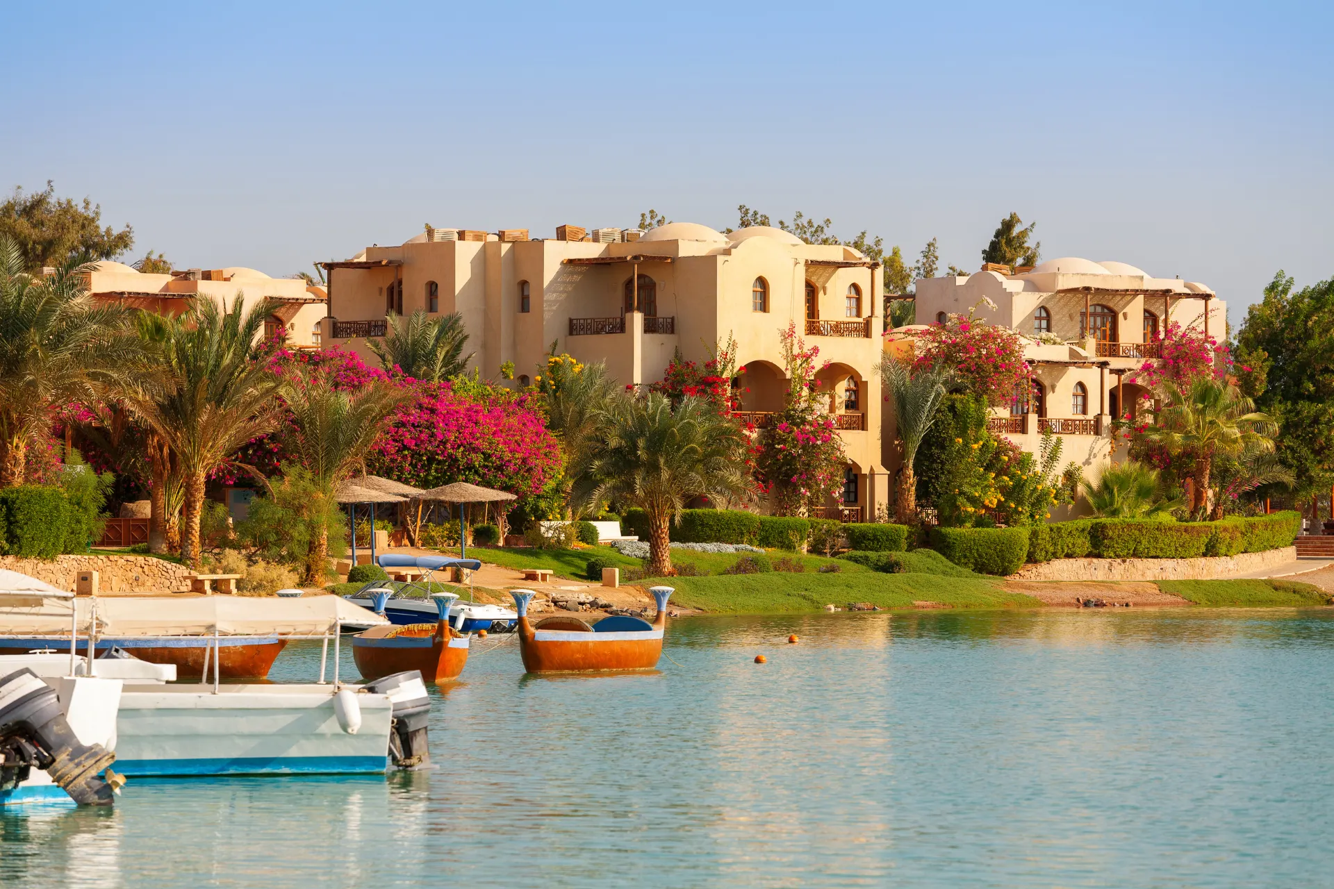 El Gouna met bootjes op het water, witte huizen en palmbomen. De zon schijnt fel onder een warme, blauwe lucht, wat een tropische en ontspannen sfeer creëert.