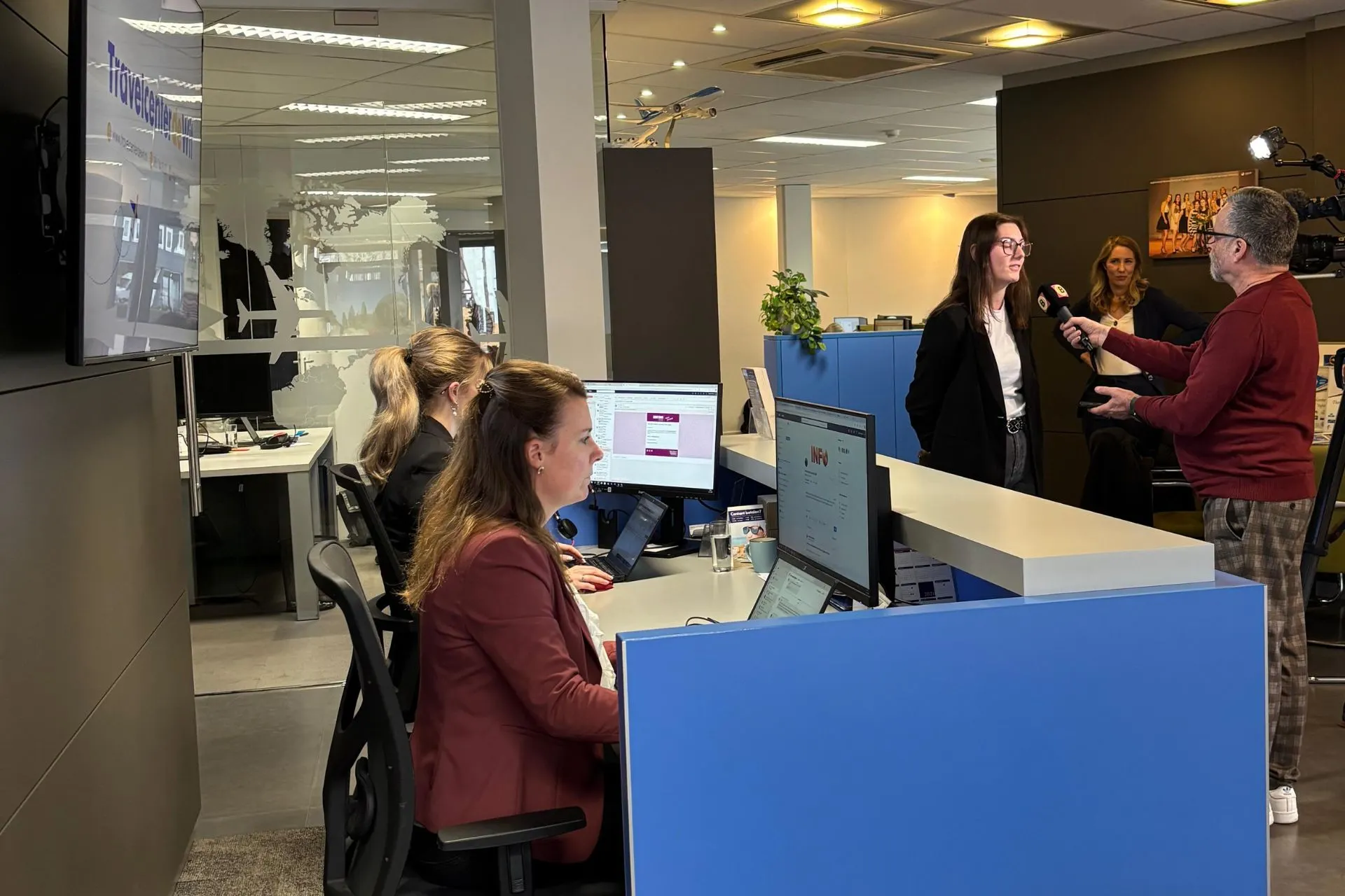 Een klant van Travelcenter de Wit wordt geïnterviewd door een journalist van Omroep Brabant bij de receptiebalie van het reisbureau. Op de achtergrond werken collega's achter hun computers, terwijl een cameraman het interview vastlegt. De moderne kantoorruimte en blauwe balie met het logo van Travelcenter de Wit zijn duidelijk zichtbaar.