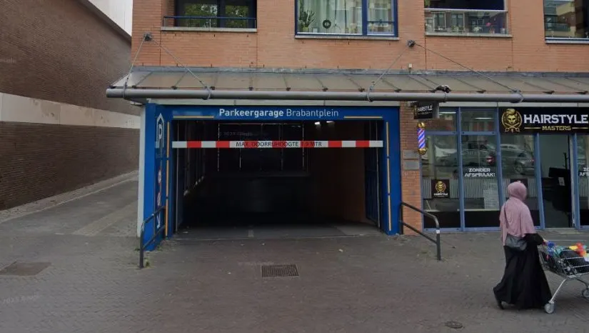 Ingang van Parkeergarage Brabantplein in Uden, met een blauwe gevel en duidelijk bord “Parkeergarage Brabantplein” boven de ingang. Er is een rood-wit hoogtebalk voor voertuigen en een donkere tunnel naar de ondergrondse parkeerplaatsen. Rechts van de ingang bevindt zich een winkel met glazen pui en een bord “Haarmode”. Op de stoep lopen voetgangers langs de garage. Stedelijke omgeving met bestrating en zijstraat zichtbaar.
