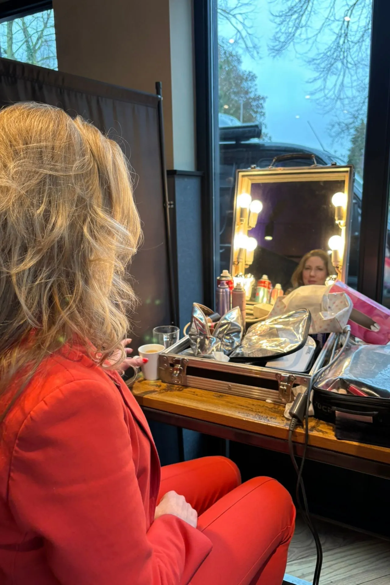 Persoon in een opvallend rood pak zit aan een tafel bij een make-up station met professionele visagiespullen, spiegels met lampen en haarproducten. Op de tafel staan een koffiekopje, sprays en een open beautycase met zilverkleurige accessoires. De setting bevindt zich bij een groot raam met daglicht en een sfeervolle achtergrond, ideaal voor voorbereidingen tijdens een fotoshoot, tv-opname of evenement. Perfect voorbeeld van backstage styling en visagie in een moderne omgeving. Omroep Brabant met Michelle de Wit en Cesar Zuiderwijk