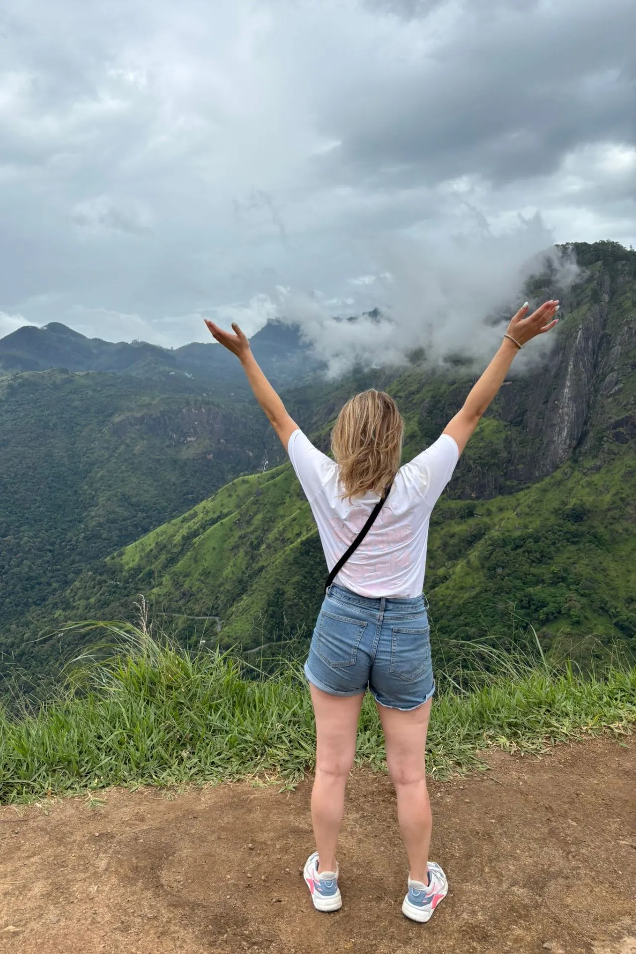 Een vrouw met opgeheven armen staat op de rand van Ella Rock en kijkt uit over de mistige bergen en groene valleien van Sri Lanka. De opgetrokken handen en weidse blik symboliseren vrijheid, verwondering en het overweldigende gevoel dat dit uitzicht biedt.