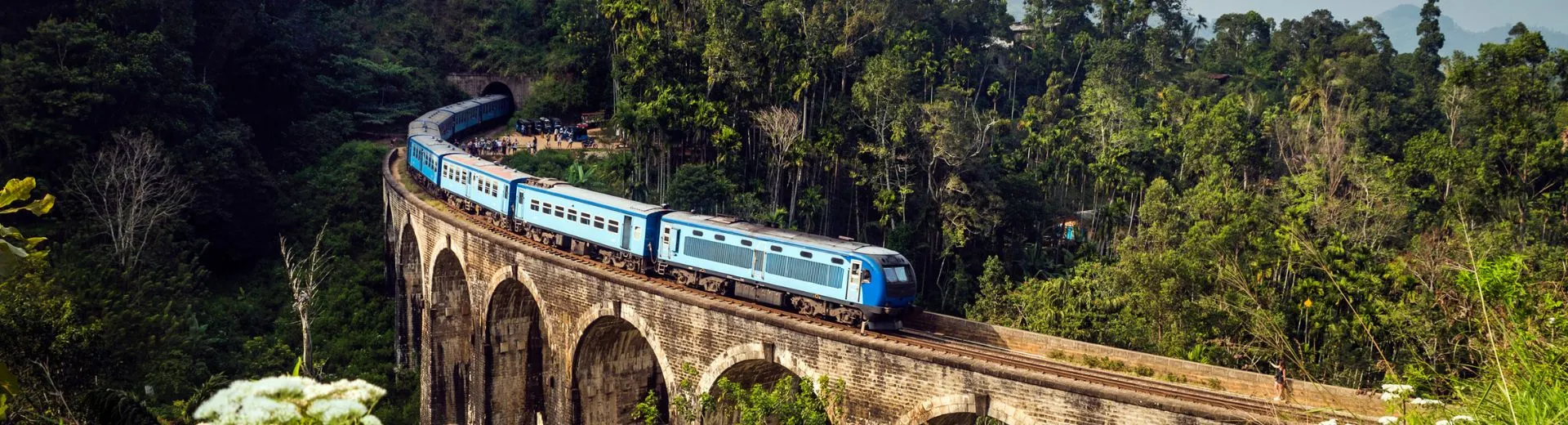 Iconische blauwe trein in Sri Lanka rijdt over de beroemde Nine Arches Bridge bij Ella, omgeven door weelderige groene jungle en berglandschap. De trein kronkelt elegant over het stenen viaduct terwijl reizigers en toeschouwers zich verzamelen bij de ingang van de tunnel. Een betoverend beeld dat de schoonheid en het avontuur van treinreizen door Sri Lanka perfect samenvat.