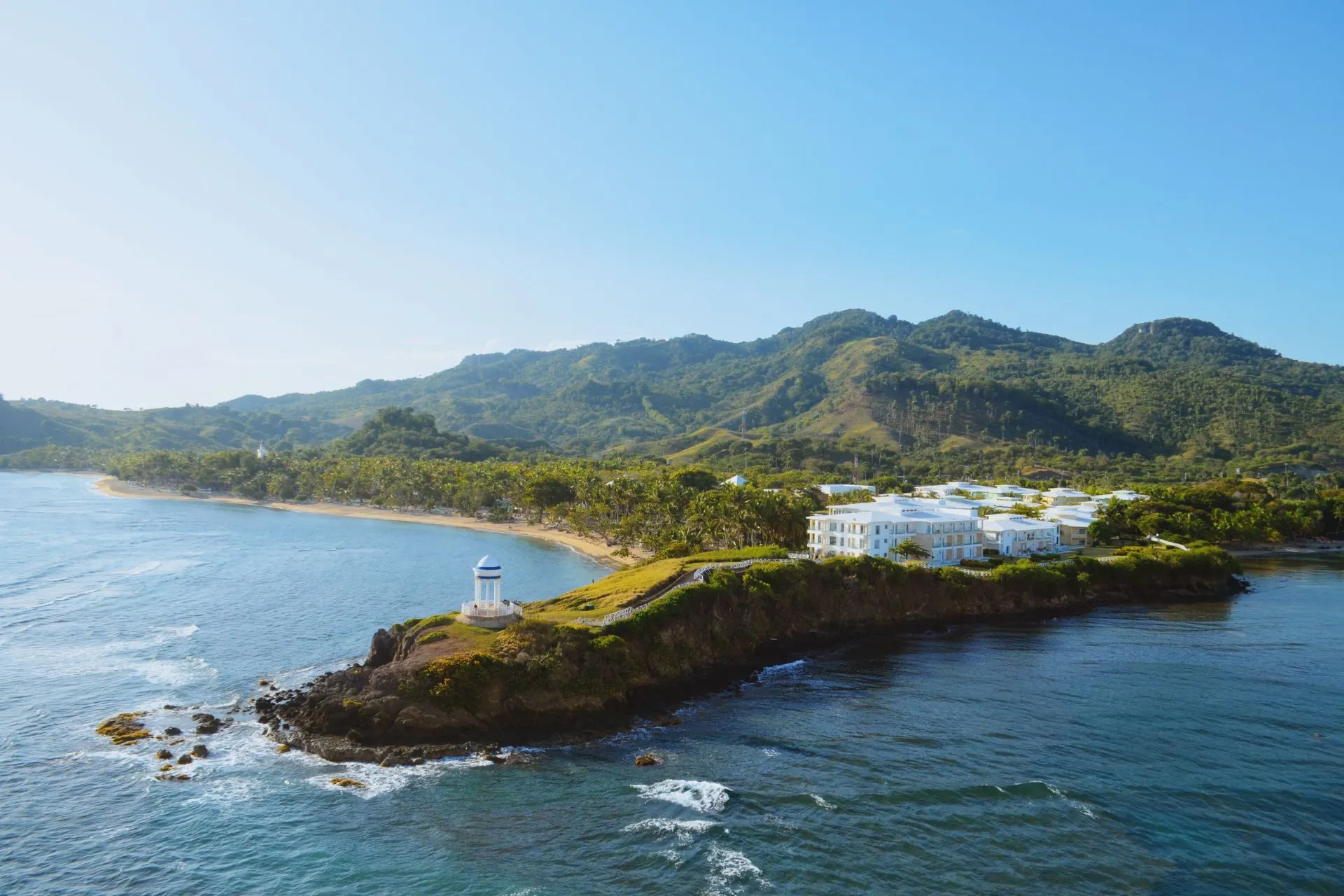 Uitzicht op de kust van Puerto Rico met witte gebouwen langs de zee en groene bergen op de achtergrond onder een heldere blauwe lucht.