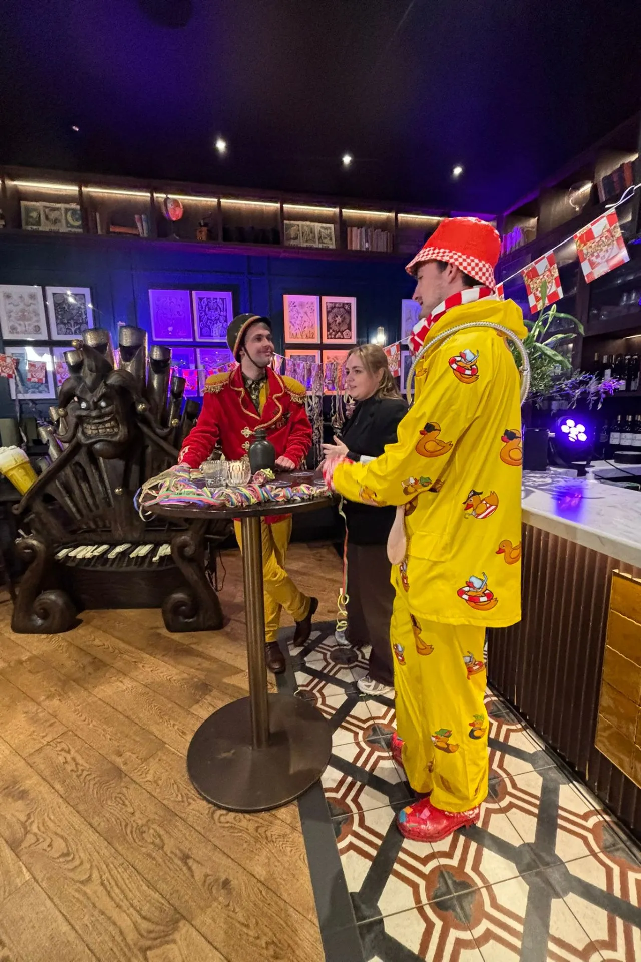 Sfeervolle carnavalsviering in een stijlvol café met kleurrijke decoraties en opvallende outfits. Een persoon draagt een felgele carnavalsoutfit met vrolijke eendjes en rode accenten, terwijl een ander een rood kostuum met gouden details draagt. Op tafel liggen kleurrijke accessoires en snoep, omringd door een uniek interieur met kunstwerken en een opvallende houten stoel. Perfecte setting voor carnaval, feestelijke borrels en Brabantse gezelligheid.