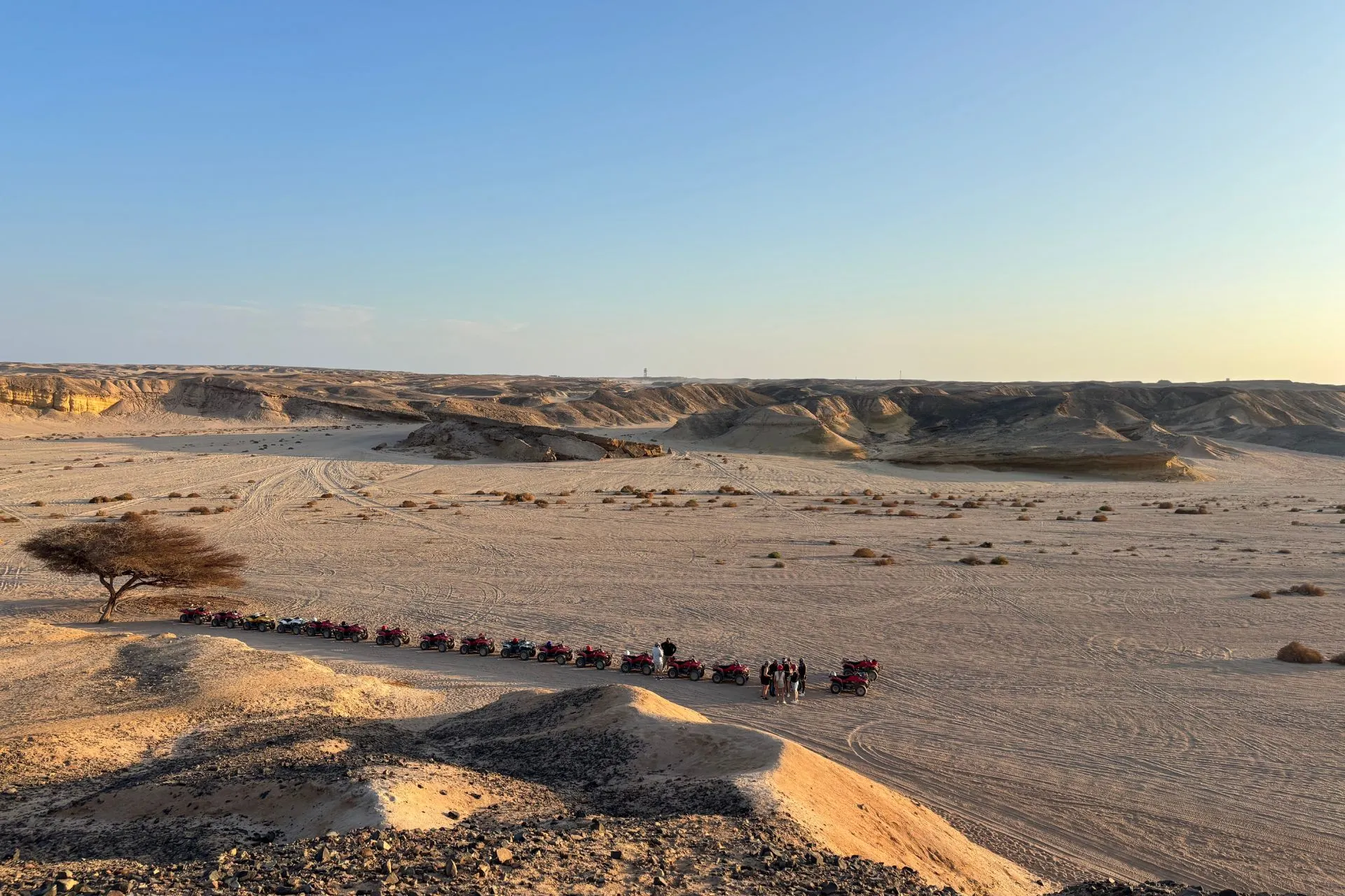 Een lange rij rode quads staat opgesteld op een zandpad in een uitgestrekt woestijnlandschap met golvende heuvels en rotsformaties onder een heldere blauwe lucht. Op de achtergrond zijn sporen zichtbaar in het zand, wat wijst op eerdere ritten door het terrein. Een enkele boom staat links in beeld, wat contrast geeft aan de droge omgeving. Dit beeld benadrukt avontuur en vrijheid, ideaal voor promotie van quadtochten, woestijnexcursies en actieve vakanties in Egypte.