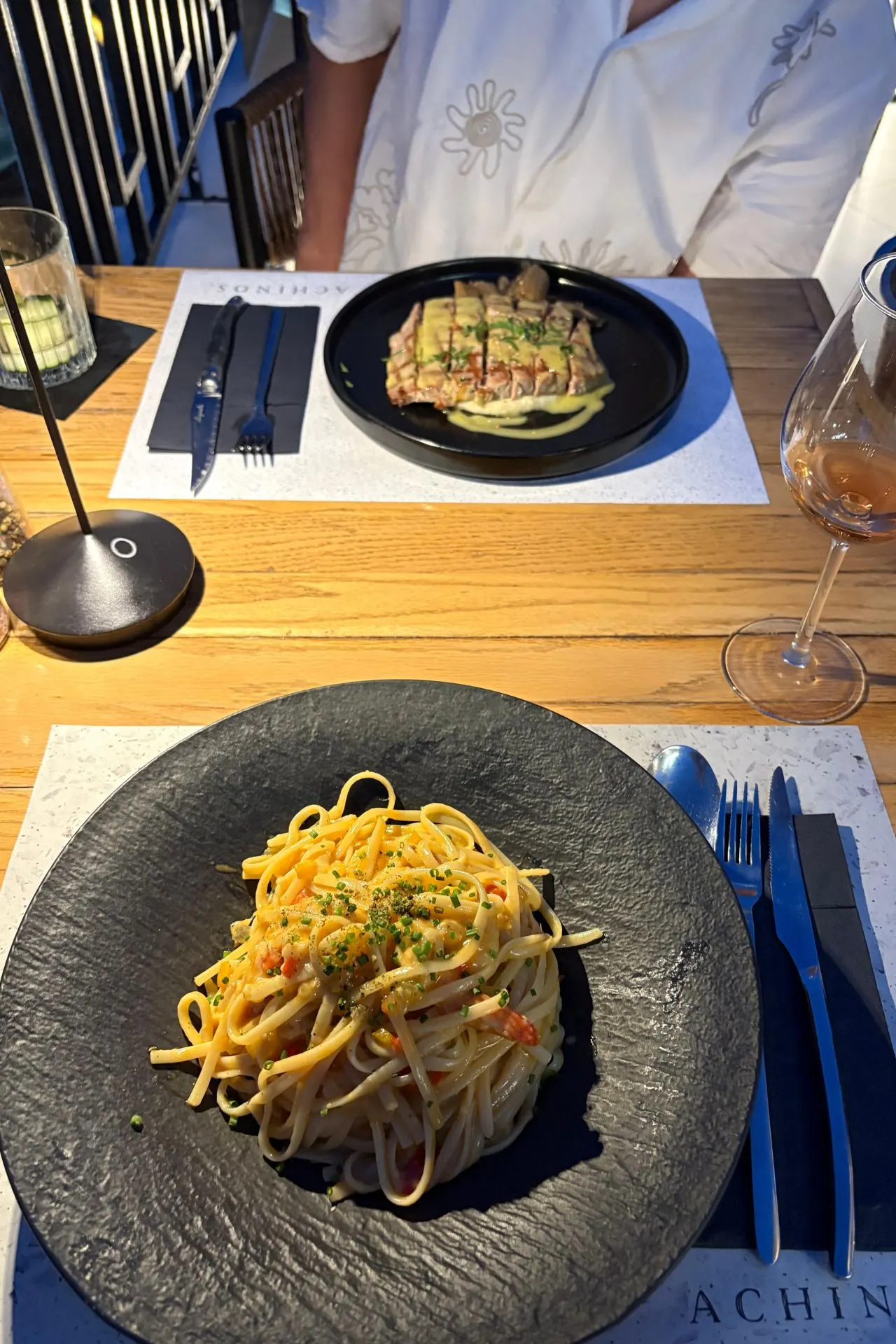 Tafel met zwarte borden met pasta en vleesgerechten in restaurant Achinos in Rethymnon, met rosé en sfeervolle verlichting.