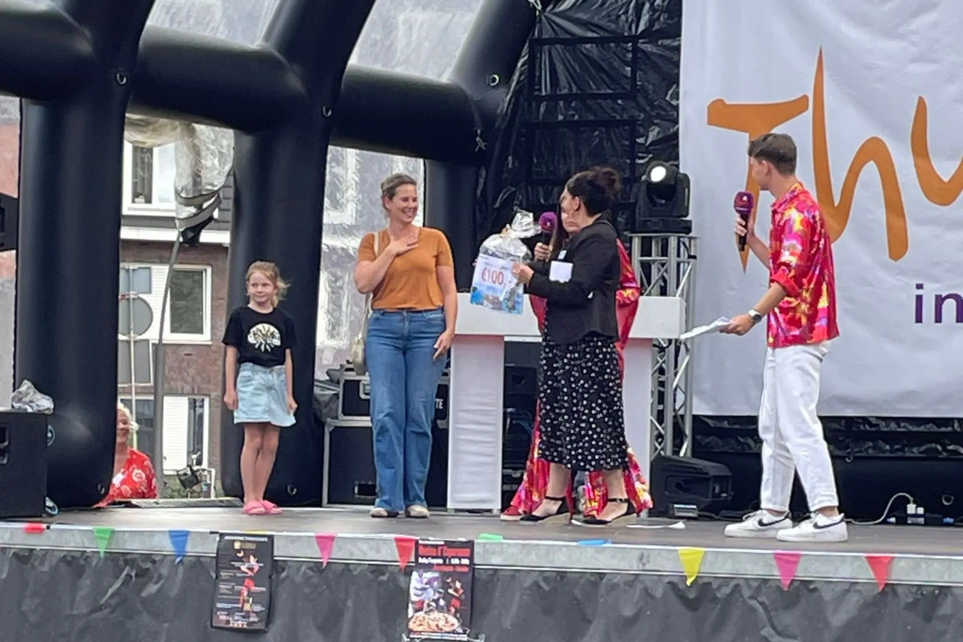 Uitreiking van een reischeque van €100 door een medewerker van Travelcenter de Wit op het podium van Thuis in Rosmalen 2025, met presentatoren en winnares met kind.