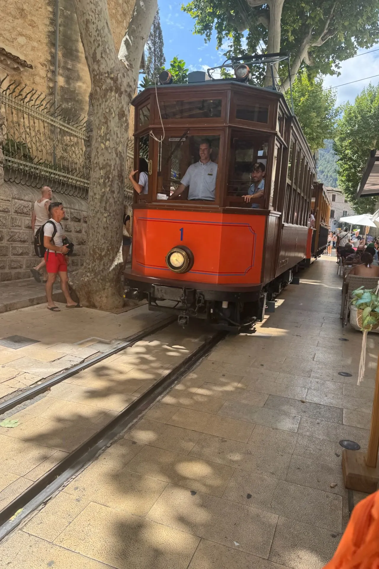 Een feloranje houten tram rijdt door een smalle straat in Sóller, Mallorca, terwijl toeristen langs de kant kijken en foto’s maken.