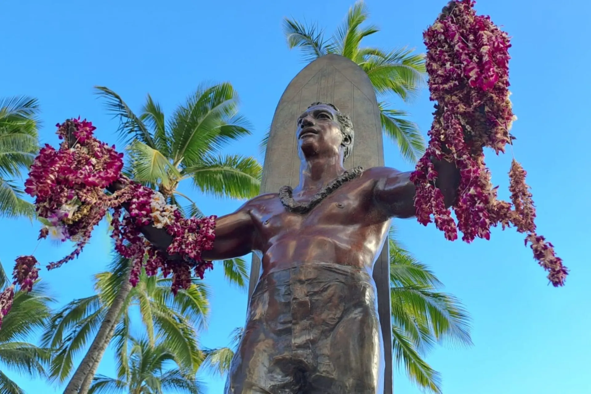 Bronzen standbeeld van Duke Kahanamoku, bekend als de vader van het moderne surfen, met bloemenkransen aan zijn handen, tegen een achtergrond van palmbomen en een heldere blauwe lucht in Waikiki, Hawaii.