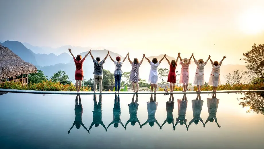Een diverse groep mensen staat hand in hand aan de rand van een infinity pool, met de armen hoog geheven in een feestelijke pose terwijl de zon ondergaat achter een schilderachtig berglandschap. De groep bestaat uit zowel mannen als vrouwen, gekleed in luchtige zomerjurken en casual outfits. Hun reflecties zijn prachtig zichtbaar in het spiegelende water van het zwembad. Op de achtergrond zijn beboste heuvels en bergen te zien in zachte blauwe en paarse tinten, wat de sfeer sereen en rustgevend maakt. Bloemen, palmen en een traditionele rieten hut zorgen voor een tropische en uitnodigende vakantiesfeer. Deze afbeelding is ideaal voor promotie van groepsreizen, retraites, vriendenvakanties of culturele vakanties op unieke locaties. Zoekwoorden: groepsreis boeken, zonvakantie in de natuur, samen reizen, vakantie met vrienden, ontspannen reizen in de bergen.