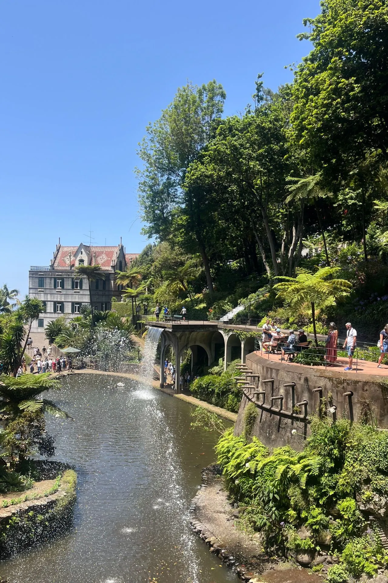 Sierlijke tuin in Funchal met fontein, brug en wandelpaden richting een elegant gebouw.
