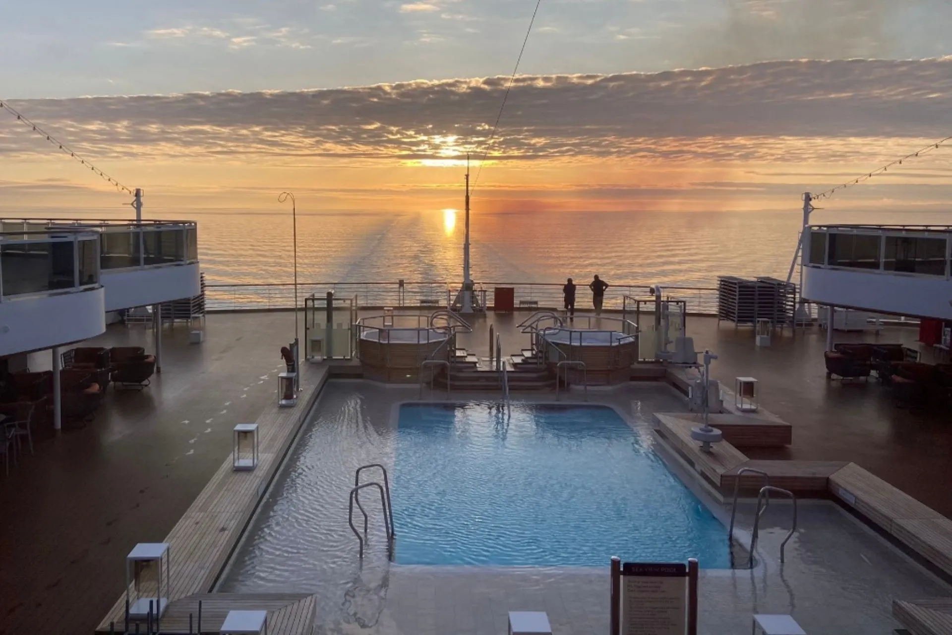 Een panoramisch uitzicht vanaf het buitendek van de Nieuw Statendam van Holland America Line, met een zwembad en jacuzzi's op de voorgrond en een prachtige zonsondergang boven de oceaan