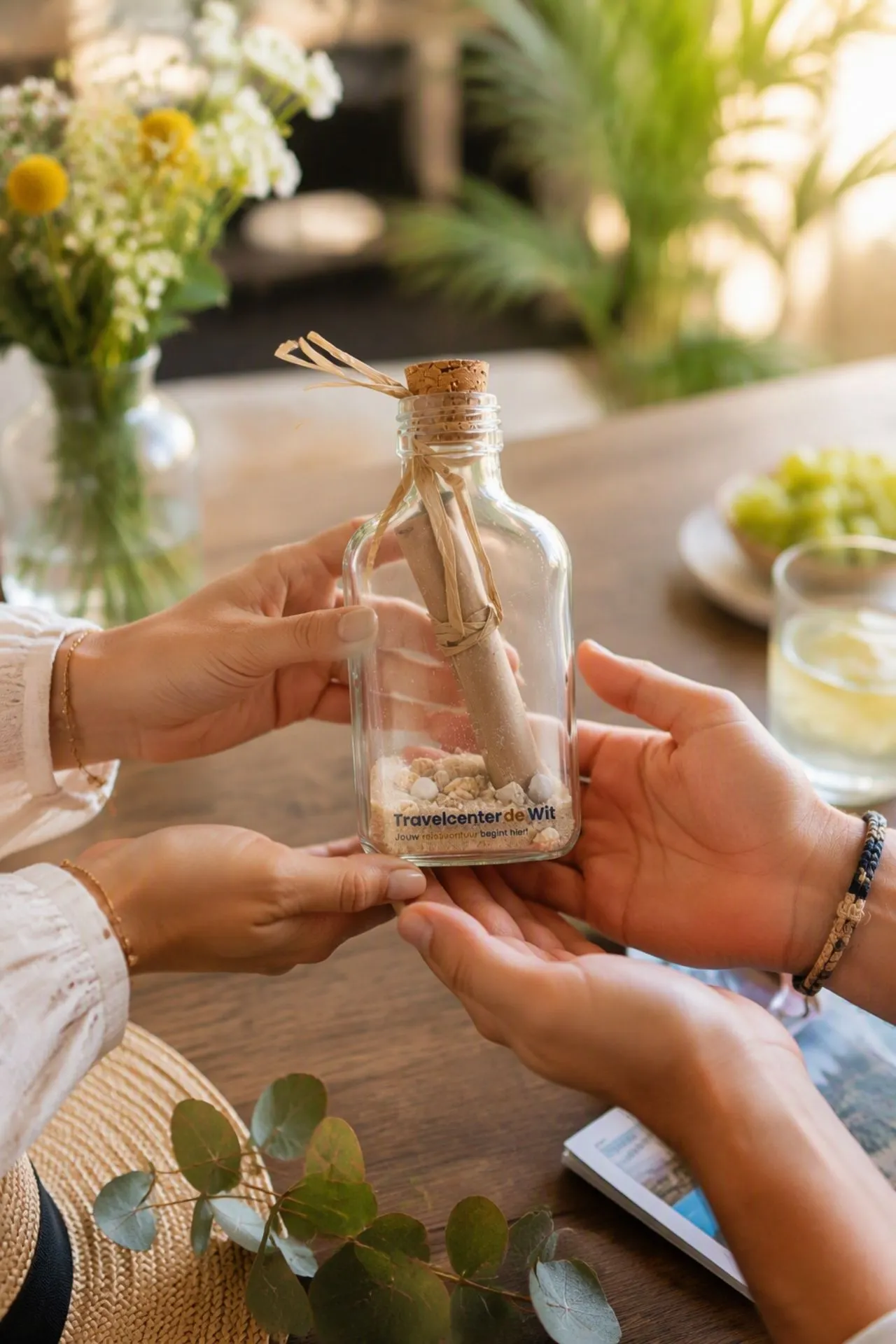 Twee personen geven een reischeque van Travelcenter de Wit aan elkaar door in een glazen fles met zand en schelpjes. Een origineel cadeau voor een verjaardag, jubileum, huwelijk of andere gelegenheid waarbij je een vakantie of reis cadeau wilt geven.