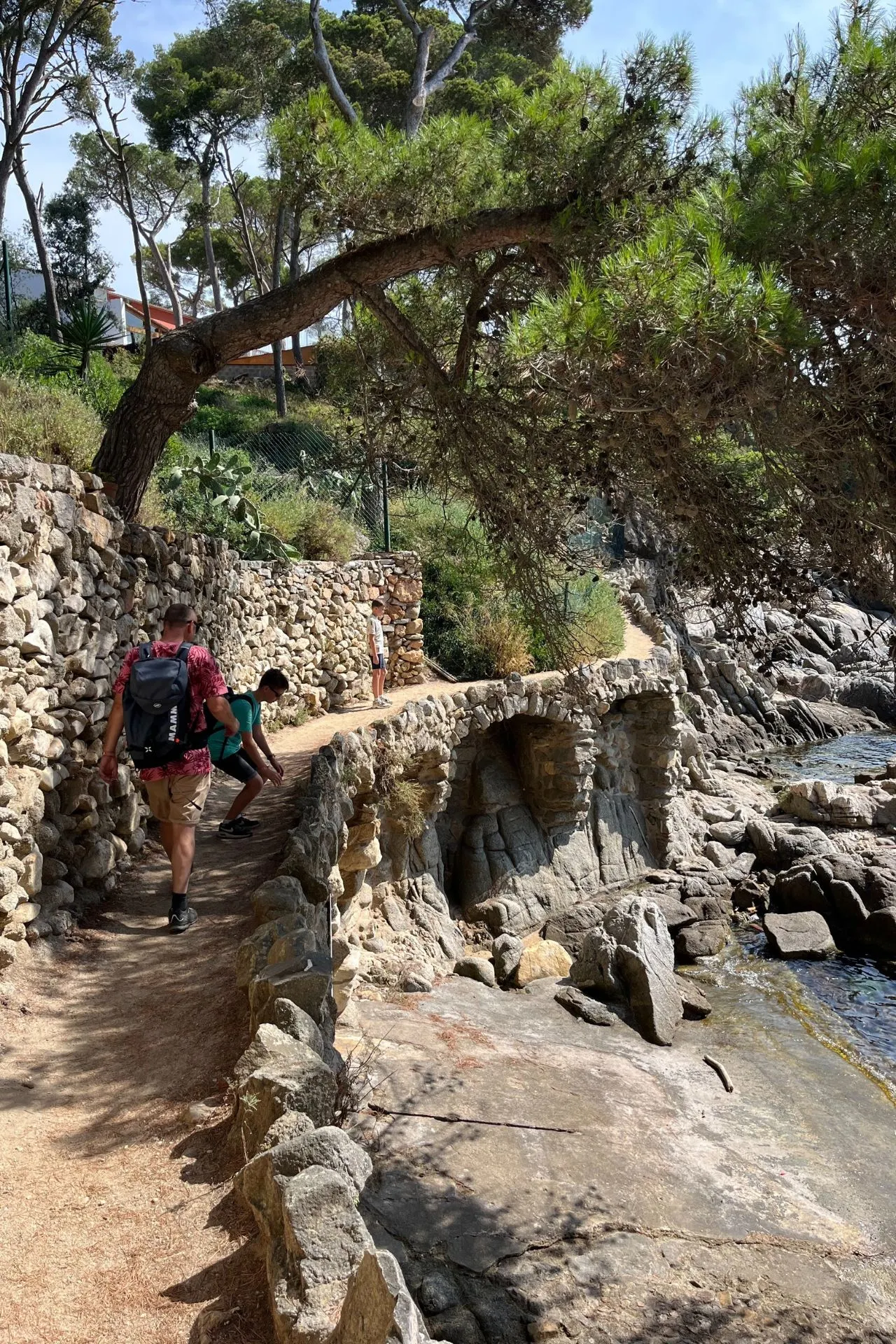 Smalle wandelroute langs een rotsachtige kust, omgeven door stenen muren en bomen. Twee wandelaars met rugzakken lopen over het pad, terwijl onder hen donkere rotsen en water zichtbaar zijn. Achtergrond gevuld met groen en zonlicht.