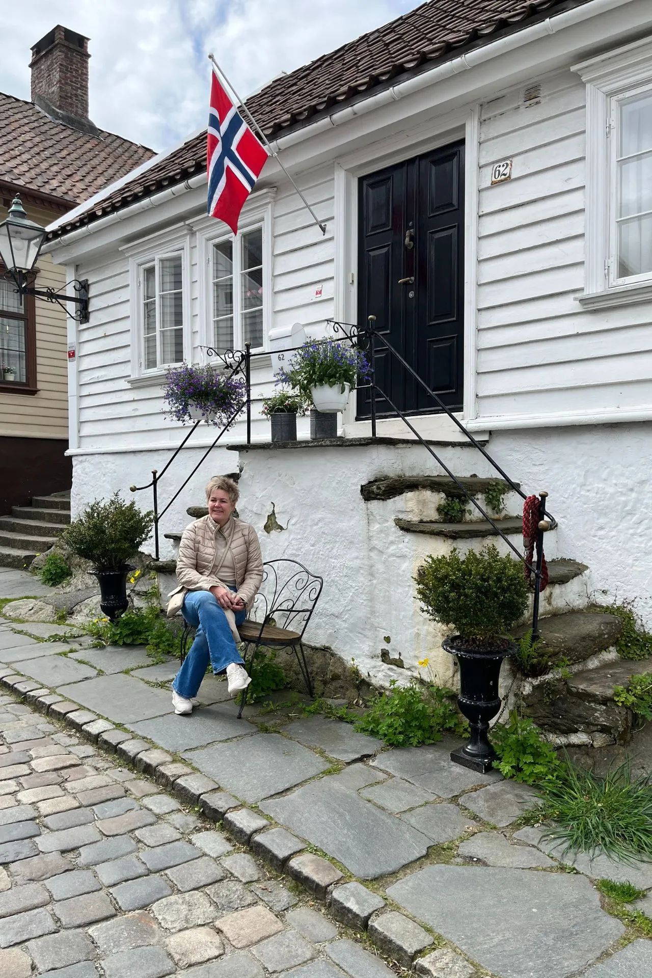 Reisspecialist Paula zittend op een bankje voor een charmant wit houten huis met een Noorse vlag en bloemen in potten, in een straat met geplaveide stenen.