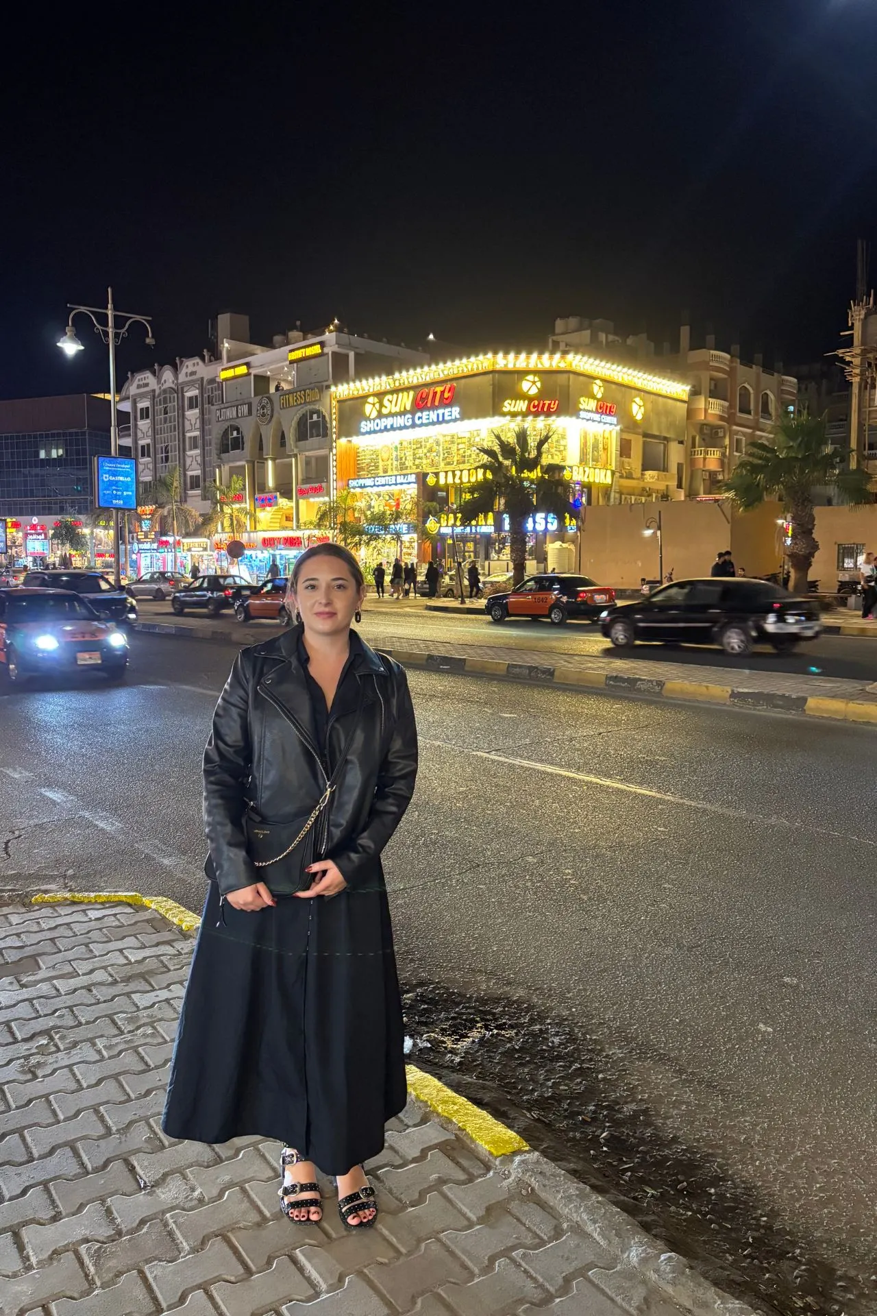 Straatbeeld in een levendige stad bij nacht met fel verlichte winkels en reclameborden, waaronder het opvallende bord “SUN GATE SHOPPING CENTER” en “SUN CITY”. Op de achtergrond zijn meerdere gebouwen met lichtdecoraties zichtbaar, terwijl auto’s over de brede weg rijden. Op de voorgrond staat een persoon in een lange zwarte jas en sandalen op een trottoir met betegeling. Dit beeld benadrukt het bruisende nachtleven en stedelijke sfeer, ideaal voor promotie van winkelen, uitgaan en stedentrips in Egypte.