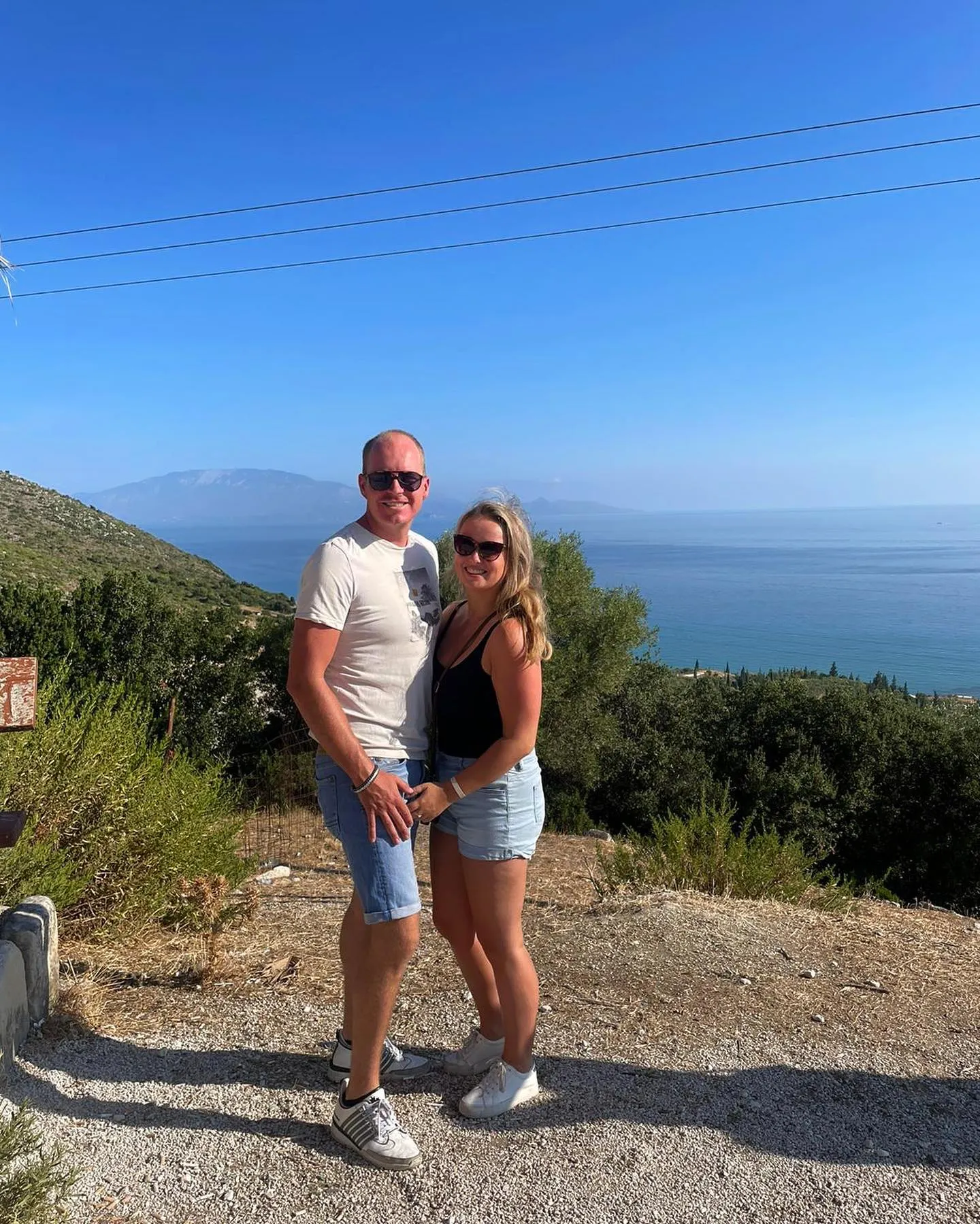 Blonde vrouw en haar partner staan op een heuvel onder een strakblauwe lucht.