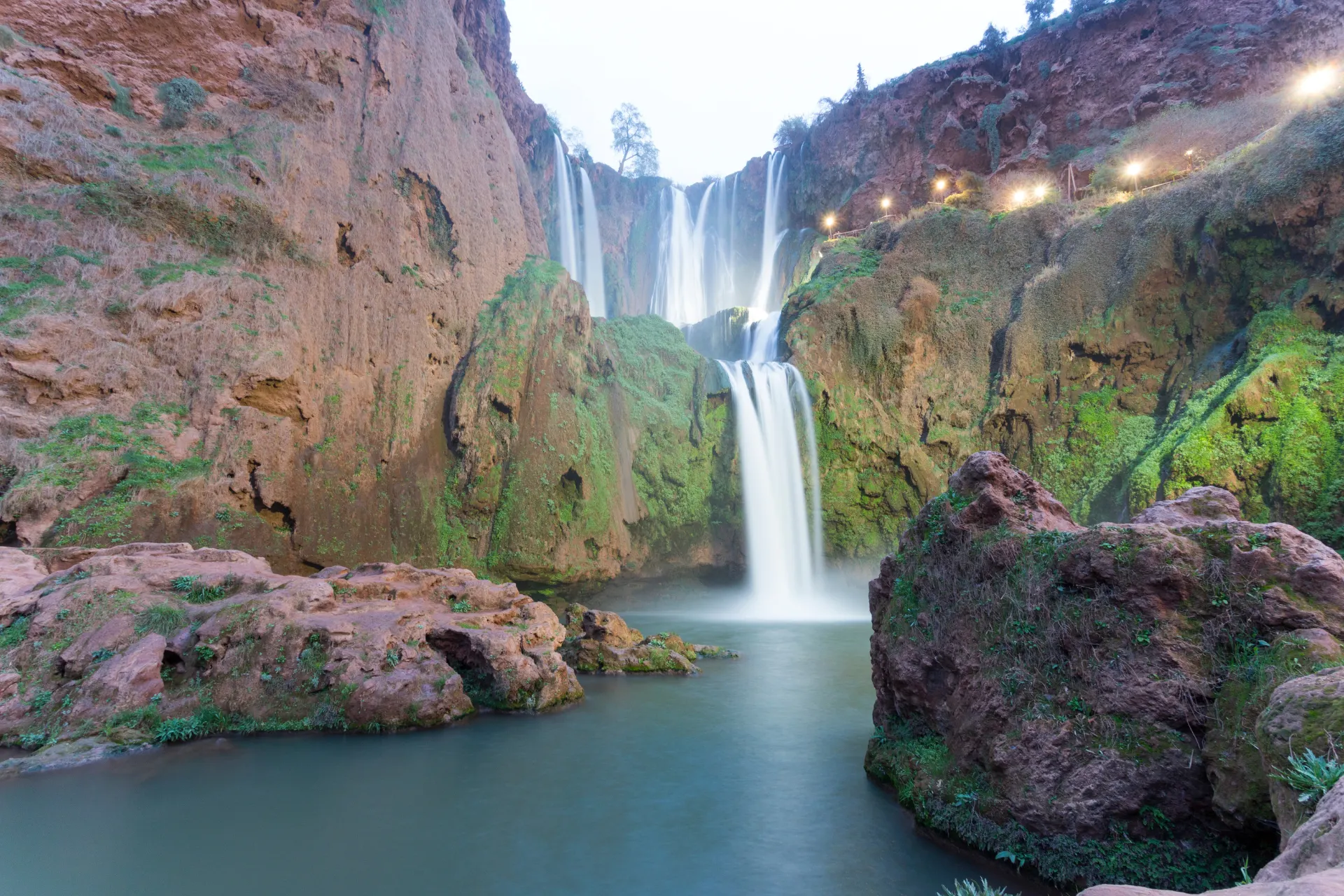 Indrukwekkende waterval die vanaf hoge rotsen in een helder bassin stort, omringd door groene mosbegroeiing en steile kliffen. Perfecte bestemming voor avontuurlijke reizen, natuurbeleving en fotografie in Marokko. Ontdek spectaculaire landschappen, verborgen valleien en serene waterpartijen in het Atlasgebergte. Ideaal voor liefhebbers van outdoor activiteiten, hiking en exotische natuur."