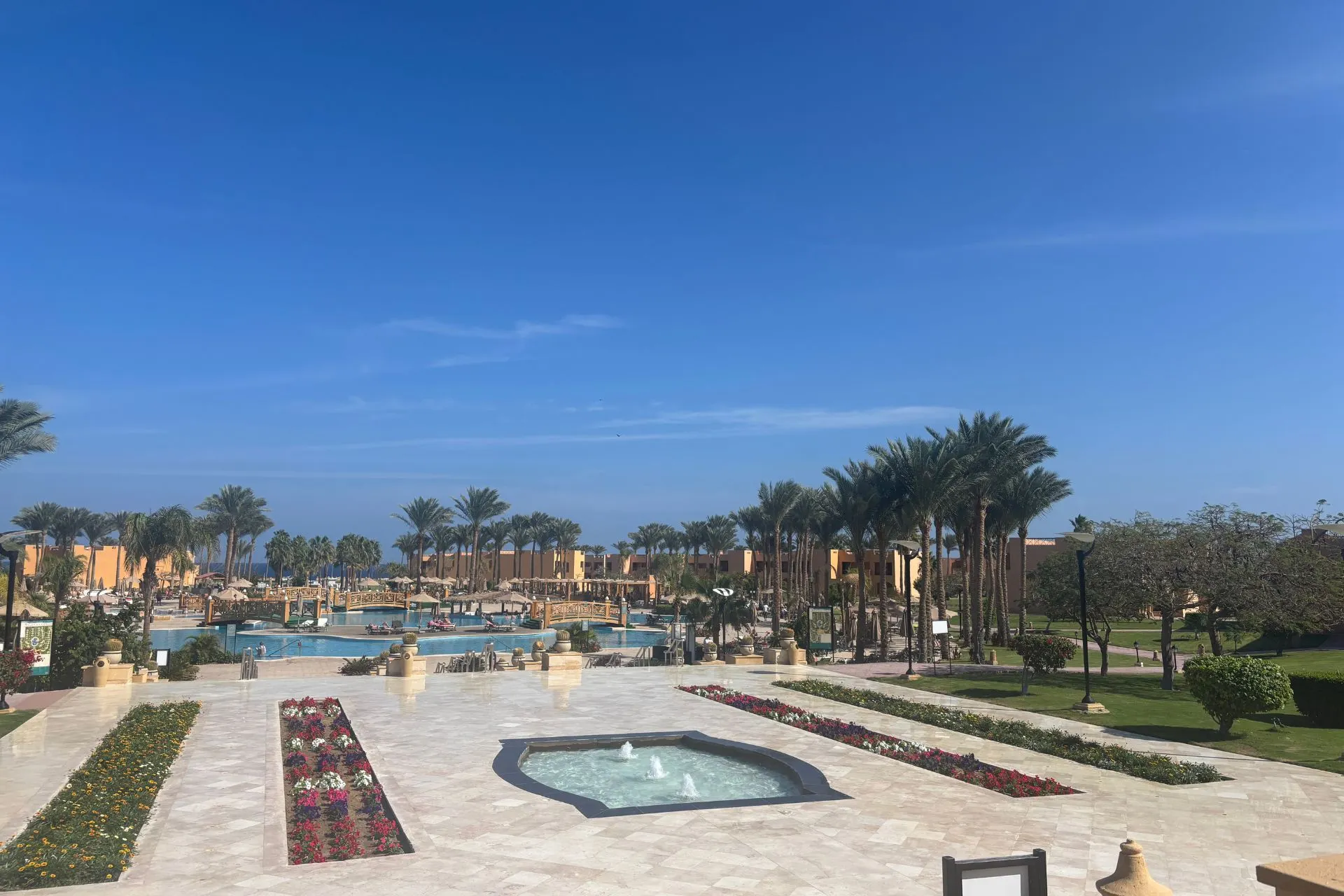 Uitzicht op een luxe resort met een groot zwembad, omringd door palmbomen en kleurrijke bloemperken. Op de voorgrond ligt een betegeld terras met een fontein en decoratieve plantenbakken. De lucht is helderblauw, wat een zonnige en ontspannen sfeer creëert. Perfect voor promotie van all-inclusive vakanties en luxe resorts in Egypte.