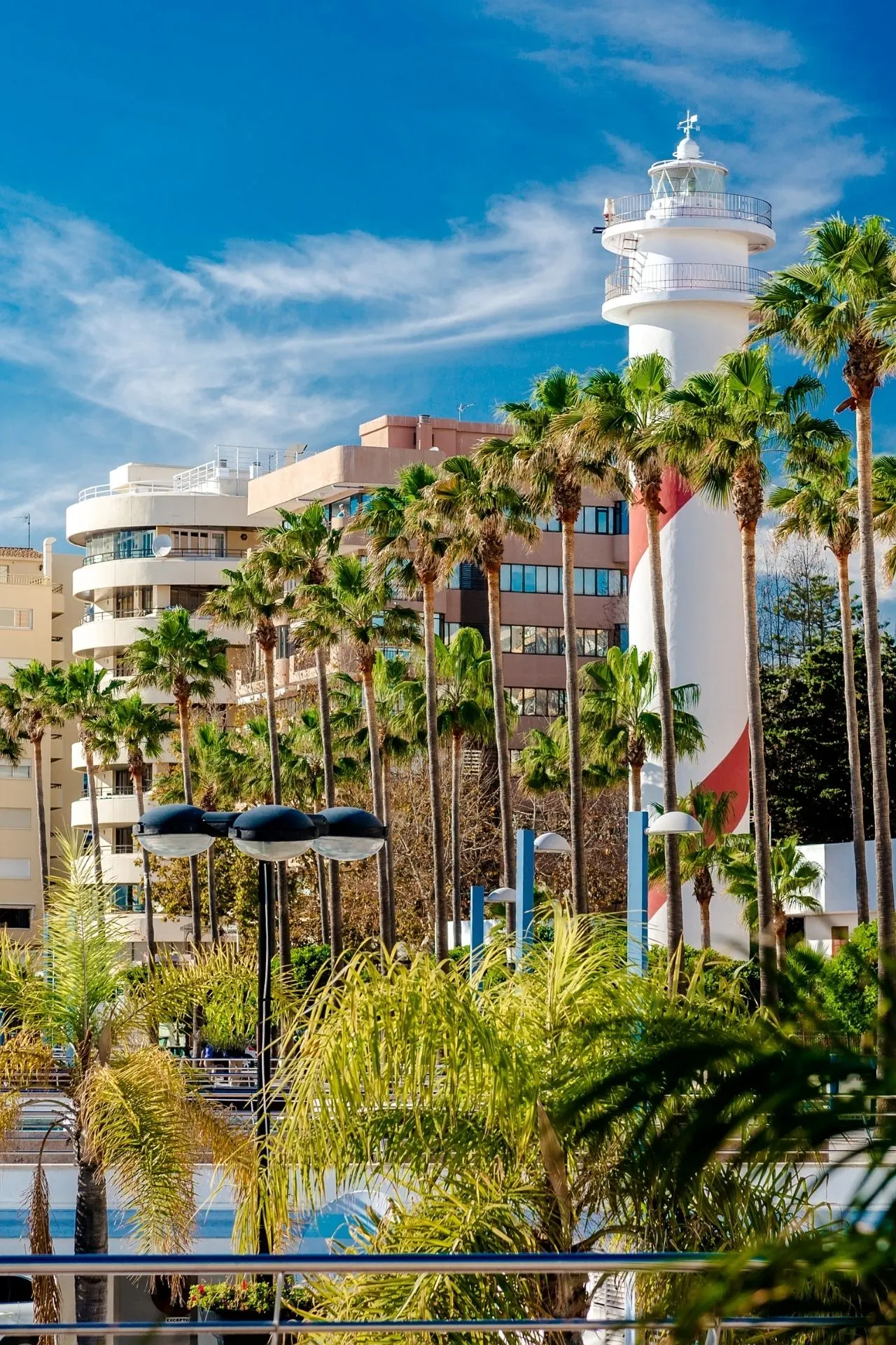 Een moderne kustomgeving met een witte vuurtoren die boven talloze slanke palmbomen uitsteekt, tegen een helderblauwe lucht met zachte wolken. Op de achtergrond staan lichte, ronde en hoekige appartementen die de mediterrane architectuur weerspiegelen. De speelse schaduwen van de palmen combineren met de zonnige kleuren en creëren een typisch zuidelijke sfeer die de levendige kustgebieden van Malaga in Spanje perfect weergeeft.