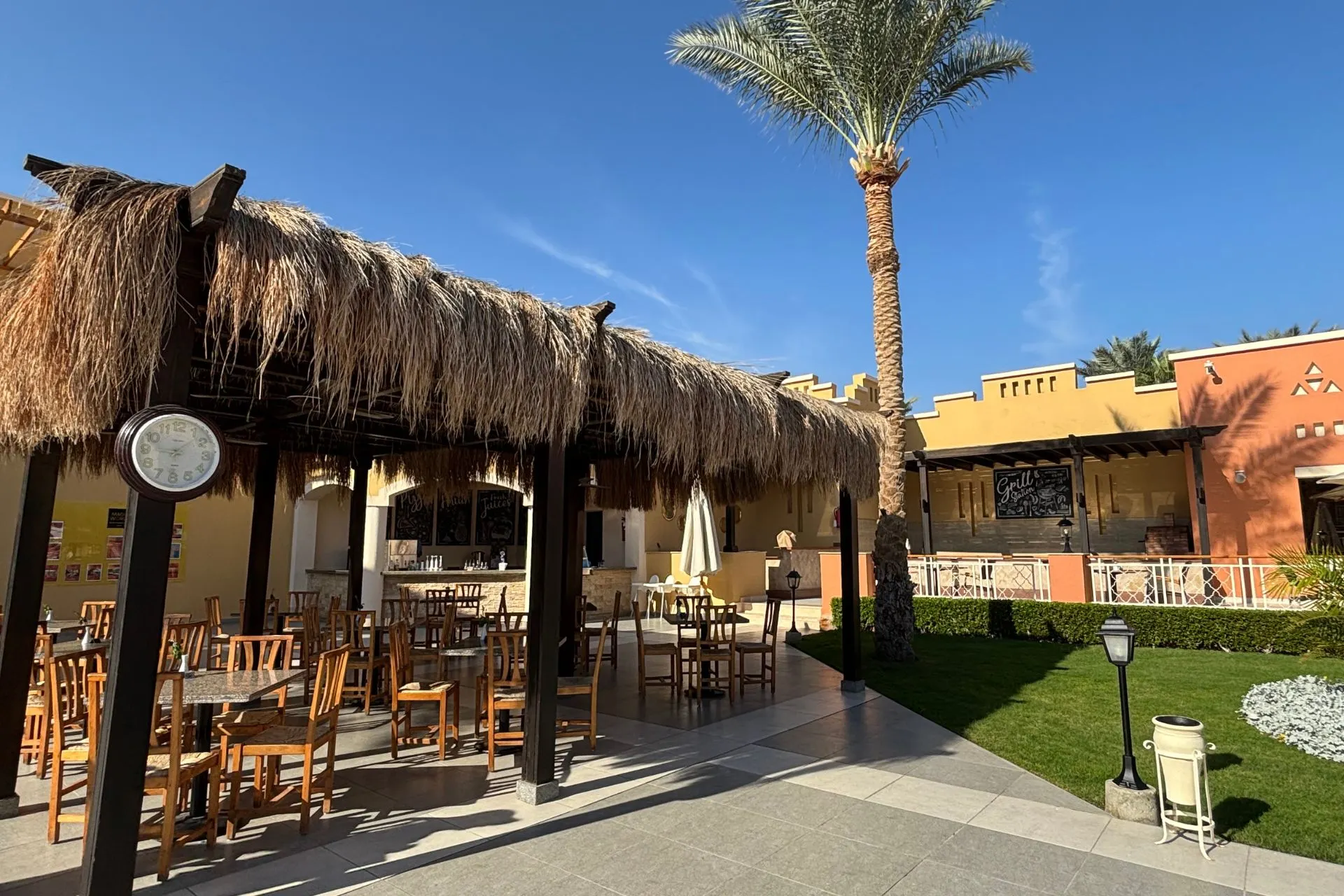 Gezellig terras met houten stoelen en tafels onder een rieten overkapping, omringd door palmbomen en groene grasvelden. Op de achtergrond staan gebouwen in warme aardetinten met decoratieve elementen. De zon schijnt fel onder een blauwe lucht, wat een uitnodigende sfeer geeft. Ideaal voor promotie van resortrestaurants en buitenactiviteiten in Egypte.