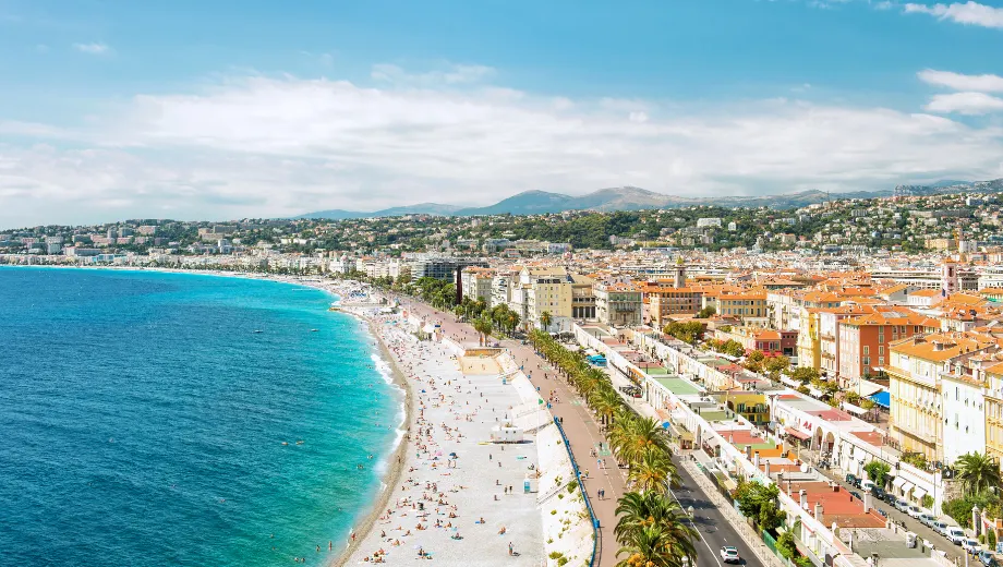 Panoramisch uitzicht op de zonnige kust van Zuid-Frankrijk met een uitgestrekte boulevard langs een helderblauwe zee. De kiezelstranden liggen vol zonaanbidders en worden omringd door palmbomen en elegante gebouwen met rode daken die de charme van de Franse Rivièra benadrukken. Op de achtergrond strekken groene heuvels zich uit onder een licht bewolkte hemel, wat zorgt voor een idyllisch vakantiegevoel. Perfect voor liefhebbers van zon, zee en cultuur die genieten van een ontspannen strandvakantie in Frankrijk.

