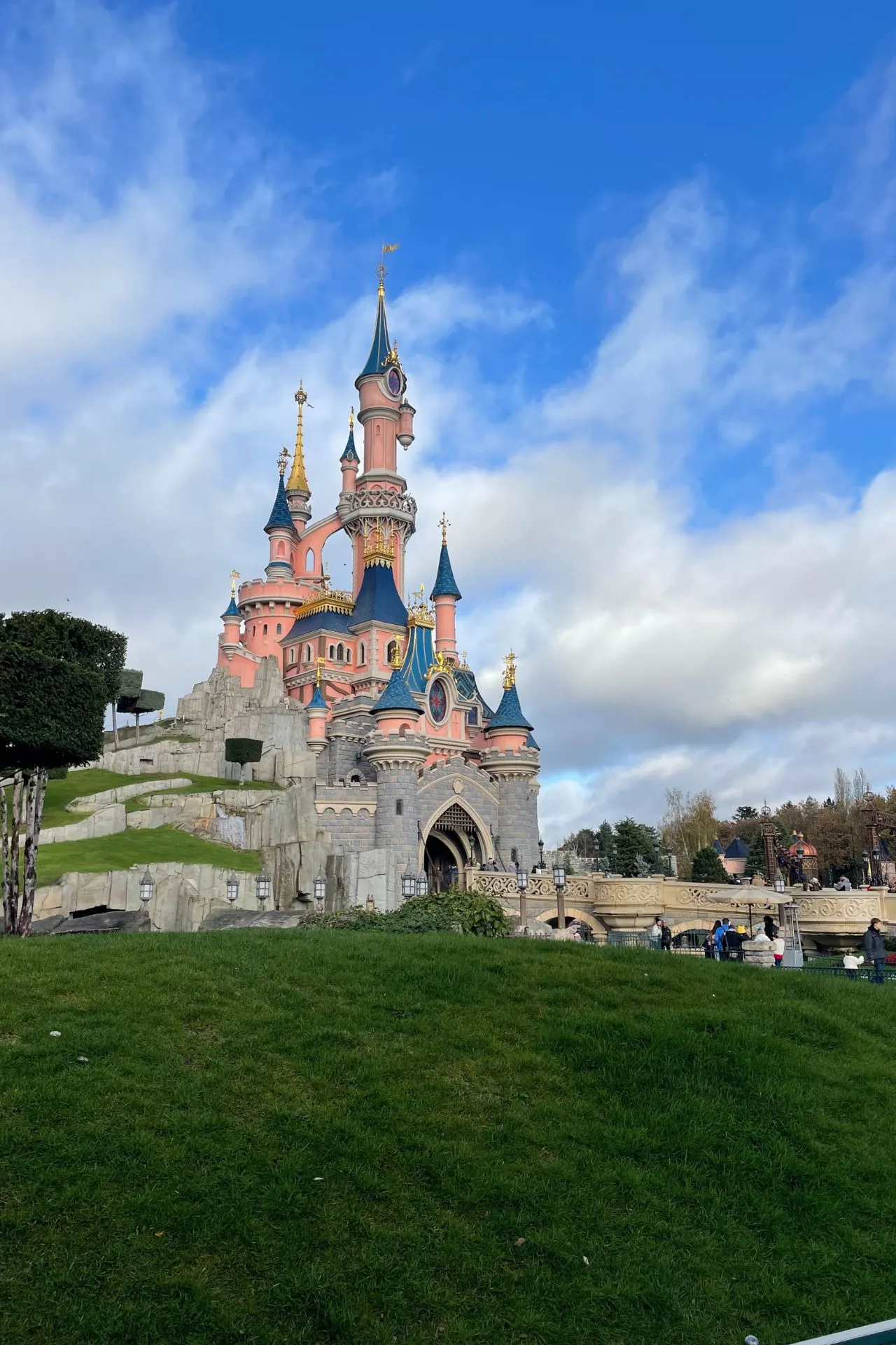 Het iconische roze kasteel van Doornroosje in Disneyland Park, Disneyland Parijs, schitterend tegen een blauwe lucht en omringd door groene heuvels. Dit sprookjesachtige kasteel is hét symbool van Disney-magie en een populaire fotospot voor bezoekers van alle leeftijden.