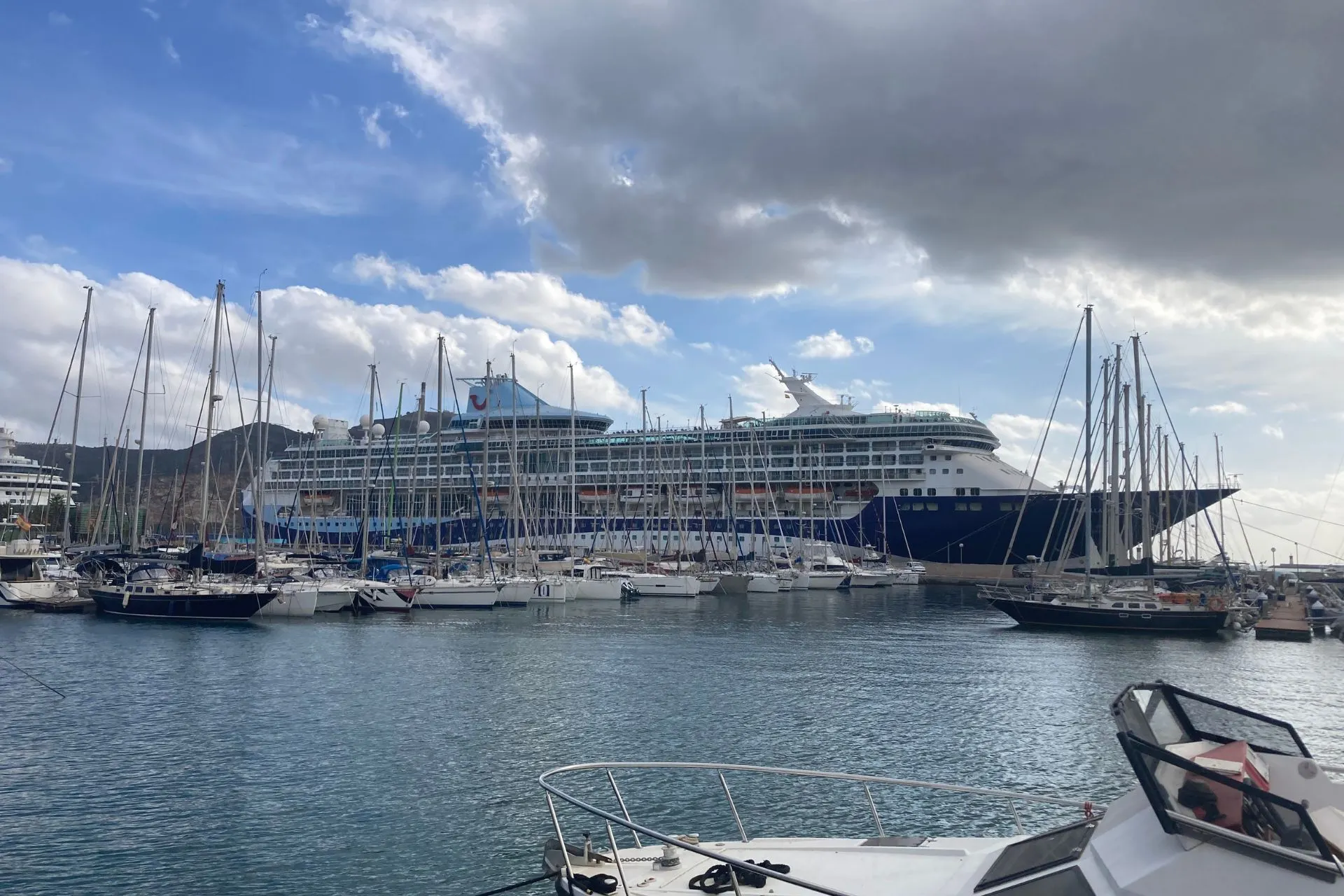 Cruiseschip aangemeerd in de haven van Cartagena, Spanje, met zeilboten op de voorgrond en een blauwe lucht met wolken.