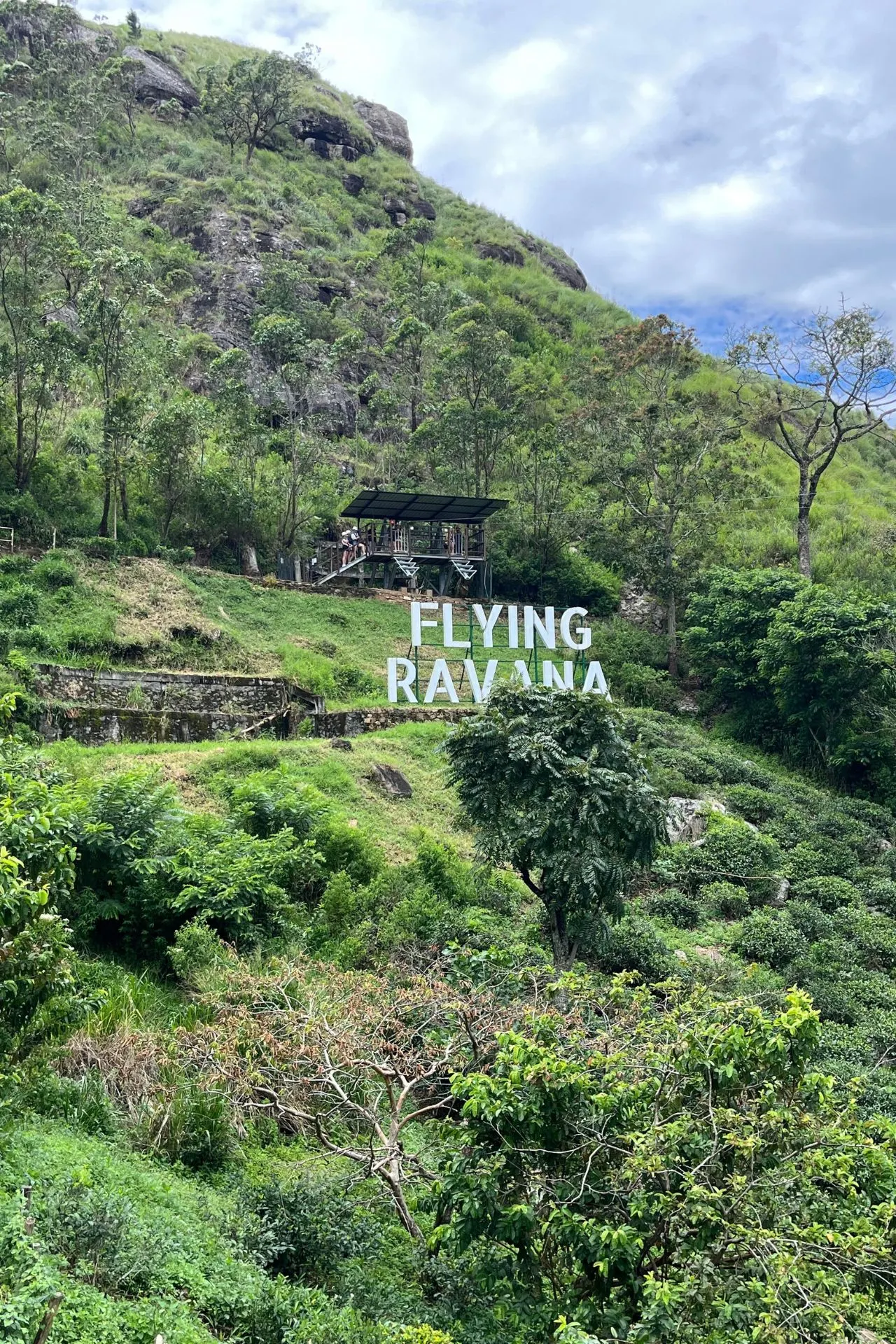 Het startplatform van de zipline bij Flying Ravana, gelegen in het weelderig groene heuvelgebied van Ella, Sri Lanka. Een groepje mensen verzamelt zich op het houten platform, met op de voorgrond het grote witte 'FLYING RAVANA'-bord dat tegen de helling staat. Een populaire hotspot voor avonturiers.