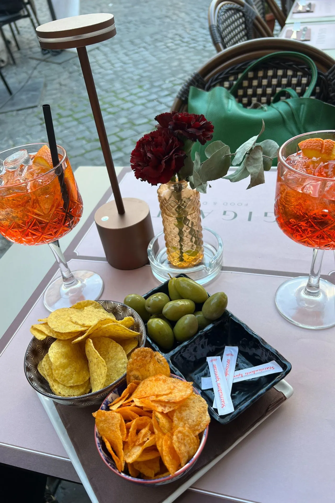 Twee glazen Aperol Spritz op een buitenterras, geserveerd met schaaltjes chips, groene olijven en servetten, op een lichtroze placemat met een kleine vaas met rode bloemen.