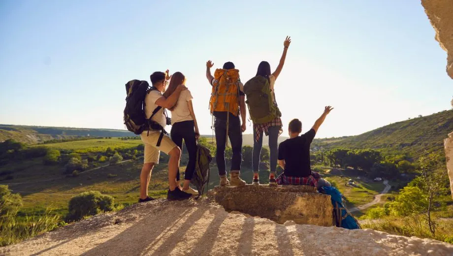 Een groep van vijf mensen met rugzakken staat op een berg en kijkt uit over een groen, heuvelachtig dal. Het landschap strekt zich uit in verschillende tinten groen onder een heldere lucht, terwijl de groep geniet van het panoramische uitzicht.