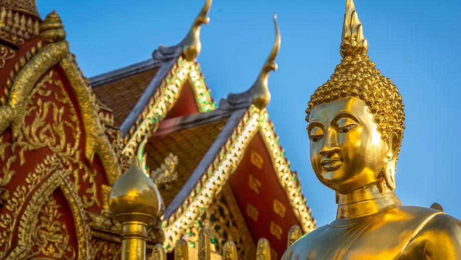 Een serene scène in Thailand met een helderblauwe lucht en een traditionele tempel met een rood en goud getrapt dak. Op de voorgrond staat een Boeddhabeeld, vredig gezeten, dat een gevoel van rust en spiritualiteit uitstraalt tegen de achtergrond van de kleurrijke architectuur.