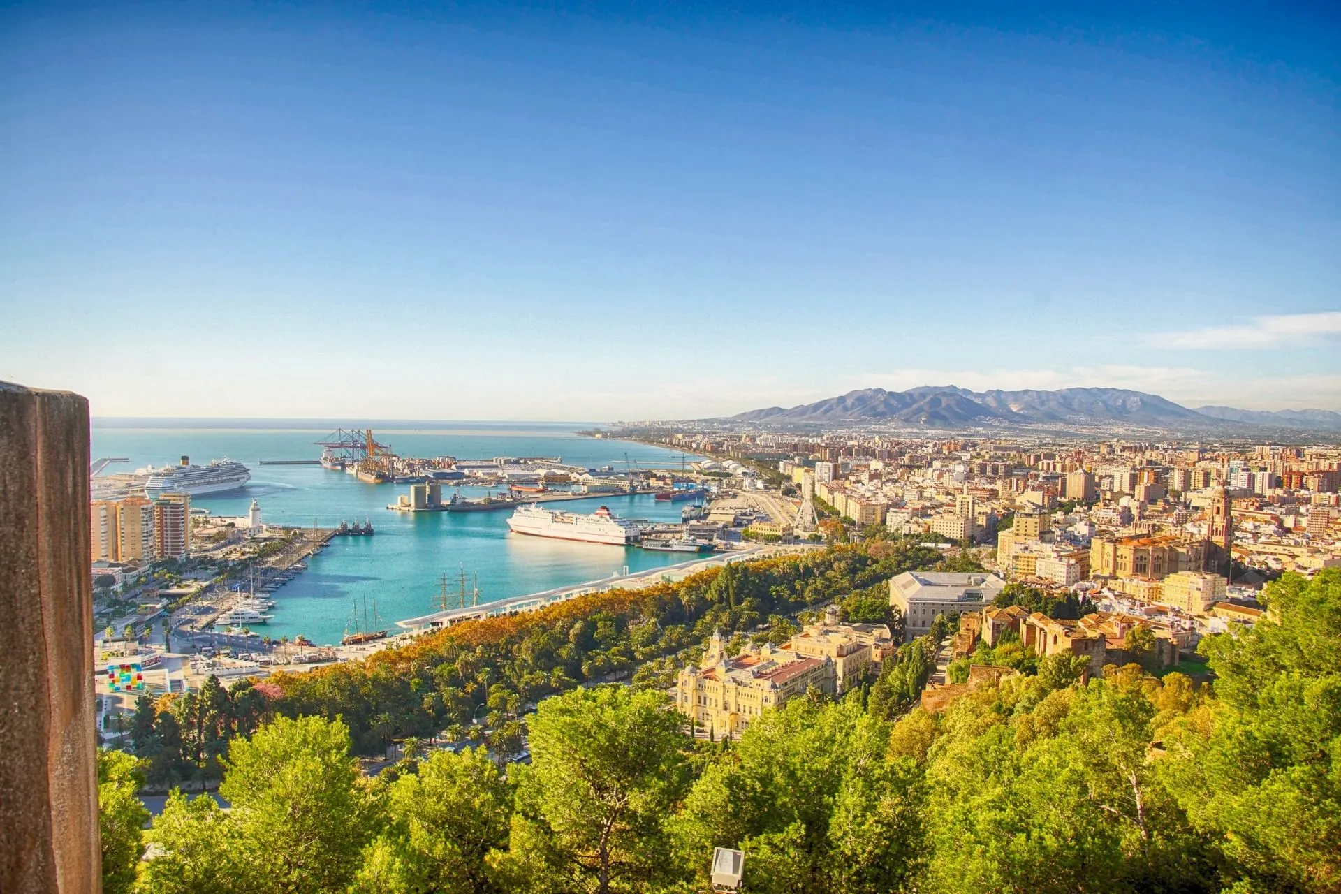 Een adembenemend panorama over de kuststad Malaga, met een helderblauwe zee, een bruisende haven vol cruiseschepen en een uitgestrekte skyline tegen de achtergrond van bergachtige landschappen. Het uitzicht toont groene parken, historische gebouwen en moderne stadsdelen die naadloos in elkaar overlopen. Deze dynamische mix van natuur, cultuur en stedelijke energie laat de veelzijdigheid van Malaga in Spanje perfect zien.