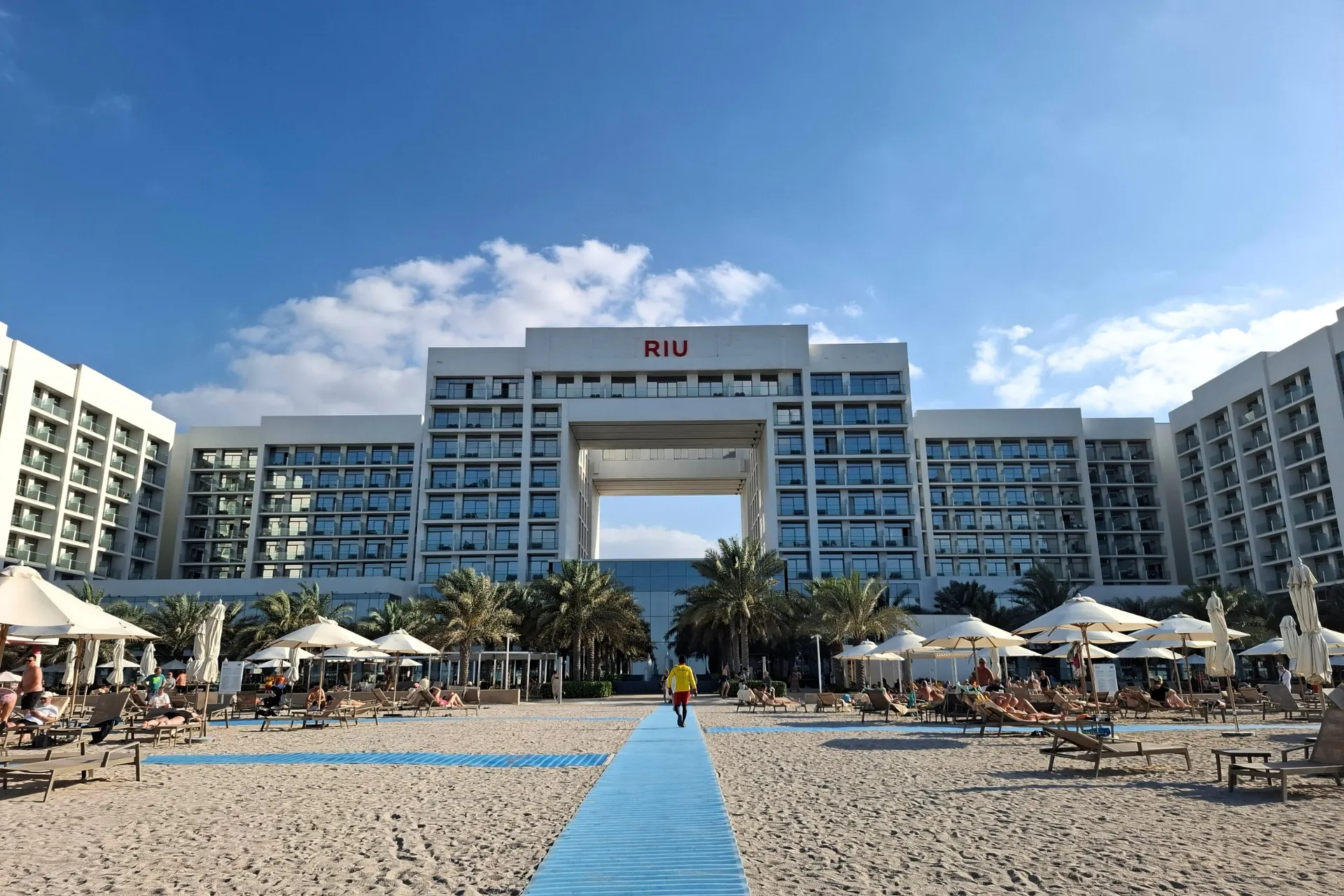 Vooraanzicht van hotel RIU in Dubai, met een blauw pad dat leidt naar het grote, rechthoekige gebouw met veel ramen, omgeven door zand.