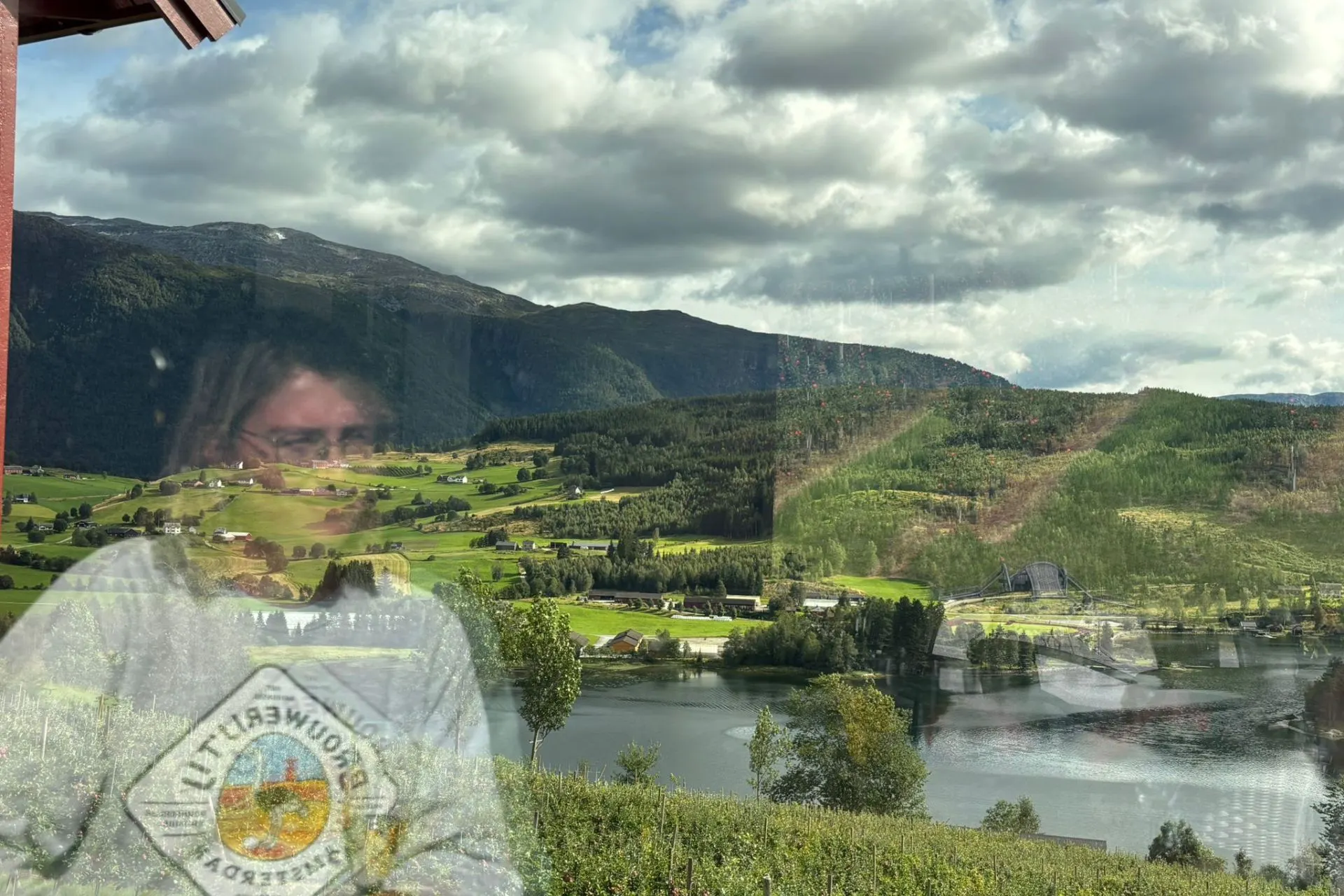 Panoramisch uitzicht op een groene vallei met verspreide boerderijen en een groot meer, omringd door beboste bergen. De lucht is deels bewolkt met zonlicht dat door de wolken schijnt.