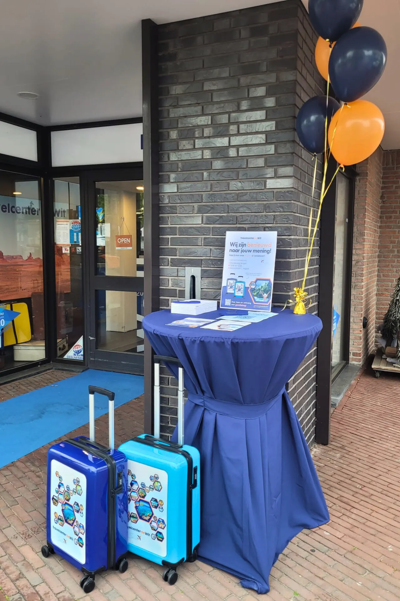 Sta tafel met blauwe tafelrok, versierd met oranje en blauwe ballonnen, in de buitenlucht voor een gezellige bijeenkomst.