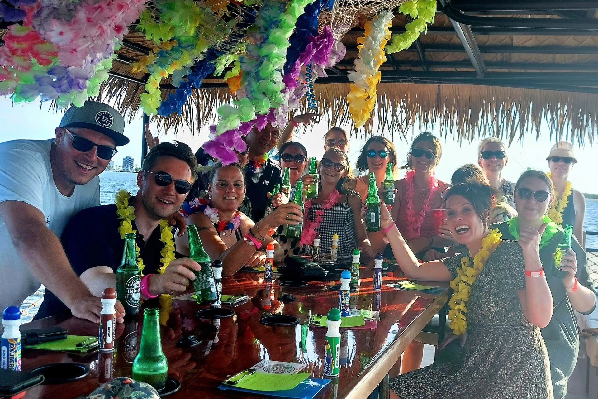 Een groep vrolijke mensen met kleurrijke bloemenkransen, genietend van drankjes aan een tropische bar op een boot.