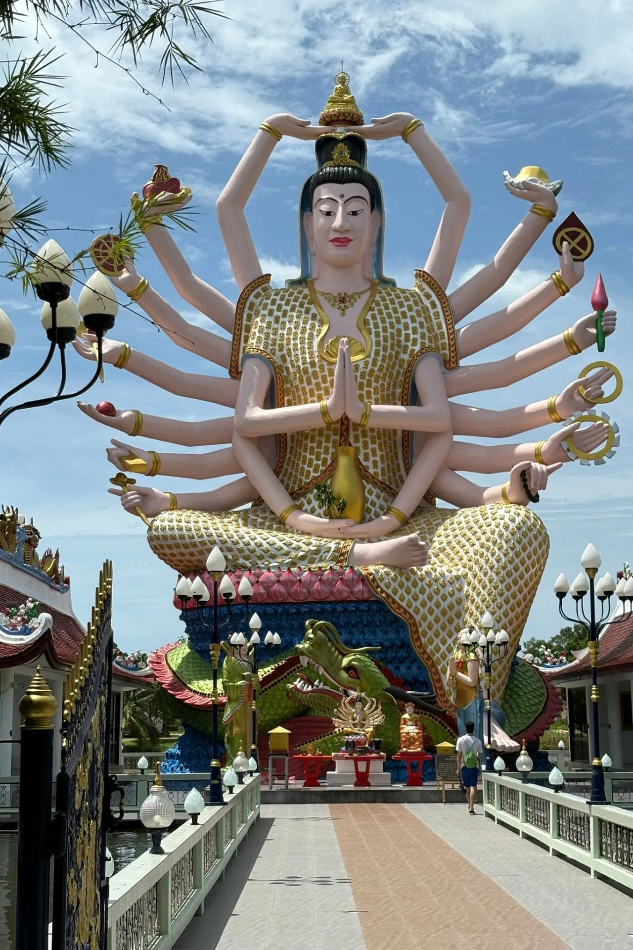 Groot kleurrijk standbeeld van de godin Guanyin met vele armen, elk met een symbolisch voorwerp, in een tempelcomplex onder een blauwe hemel.