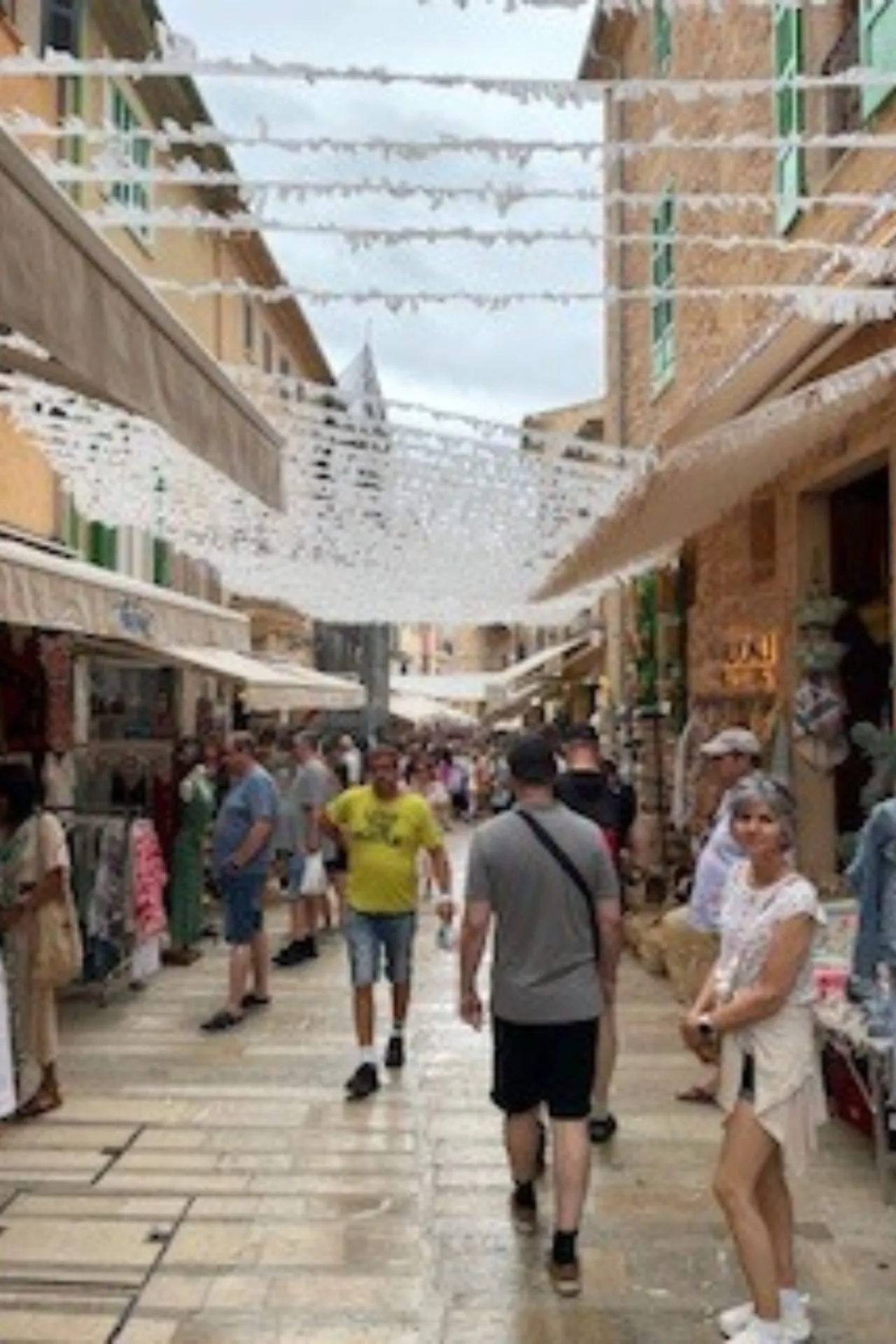 Gezellige smalle straat in een historisch centrum met sfeervolle winkels en boetieks aan beide zijden. Decoratieve witte slingers hangen boven de straat en creëren een charmante mediterrane ambiance. Mensen wandelen langs terracotta gevels en luifels, terwijl de geplaveide straat een authentieke uitstraling geeft. Perfecte bestemming voor sightseeing, winkelen en cultuurbeleving in een pittoresk dorp op Mallorca.