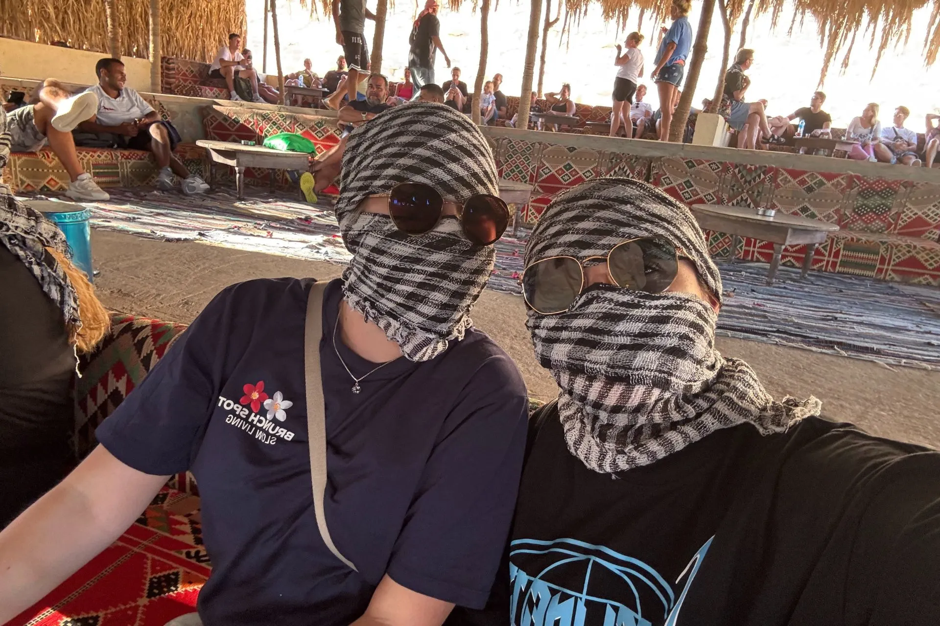 Twee personen zitten naast elkaar in een woestijnkamp onder een rieten afdak, gekleed in casual shirts en met zwart-wit geblokte sjaals om hun hoofd en gezicht gewikkeld ter bescherming tegen zand en zon. Beide dragen zonnebrillen, wat een stoere en praktische uitstraling geeft. Op de achtergrond zijn kleurrijke tapijten, lage zitplaatsen en andere mensen zichtbaar, wat een ontspannen sfeer van een woestijnexcursie benadrukt. Ideaal beeld voor promotie van woestijnsafari’s, culturele ervaringen en avontuurlijke vakanties in Egypte.
