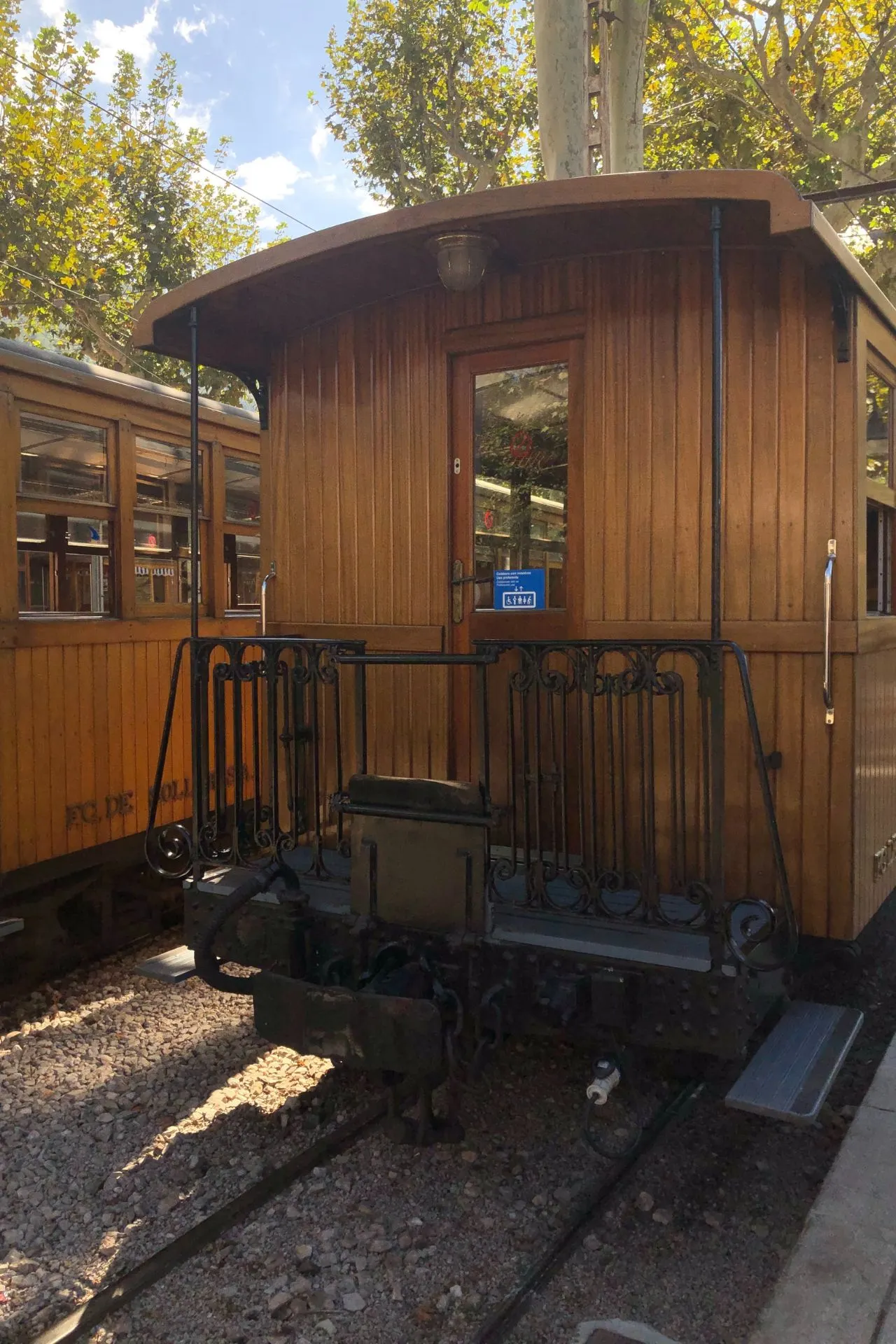Historische houten treinwagon van de beroemde Sóller-trein op Mallorca, geparkeerd op het station tussen groene bomen en onder een zonnige blauwe lucht. Deze nostalgische treinrit door de Serra de Tramuntana is een van de populairste excursies op het eiland en biedt een unieke ervaring voor reizigers die cultuur en natuur willen combineren tijdens hun Mallorca-vakantie.