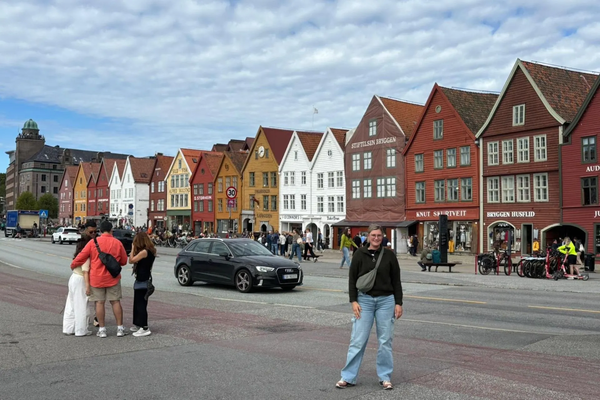 Rij kleurrijke houten huizen in traditionele stijl langs een straat in een historische wijk. Voor de huizen lopen mensen en staan fietsen, terwijl auto's op de weg rijden. De lucht is deels bewolkt.